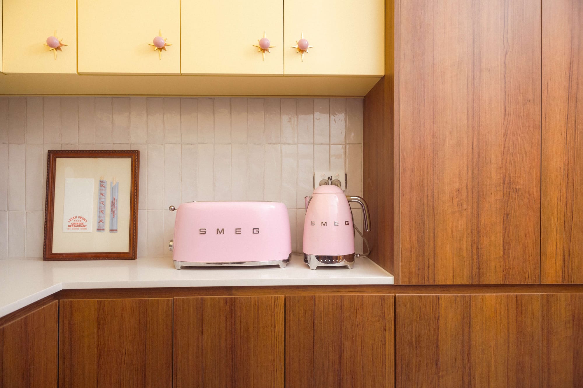 A kitchen bench detail with pale yellow overhead cabinetry fitted with starburst hardware, warm timber lower cabinets, a handmade tile splashback, and a matching pink Smeg toaster and kettle alongside a framed vintage menu.