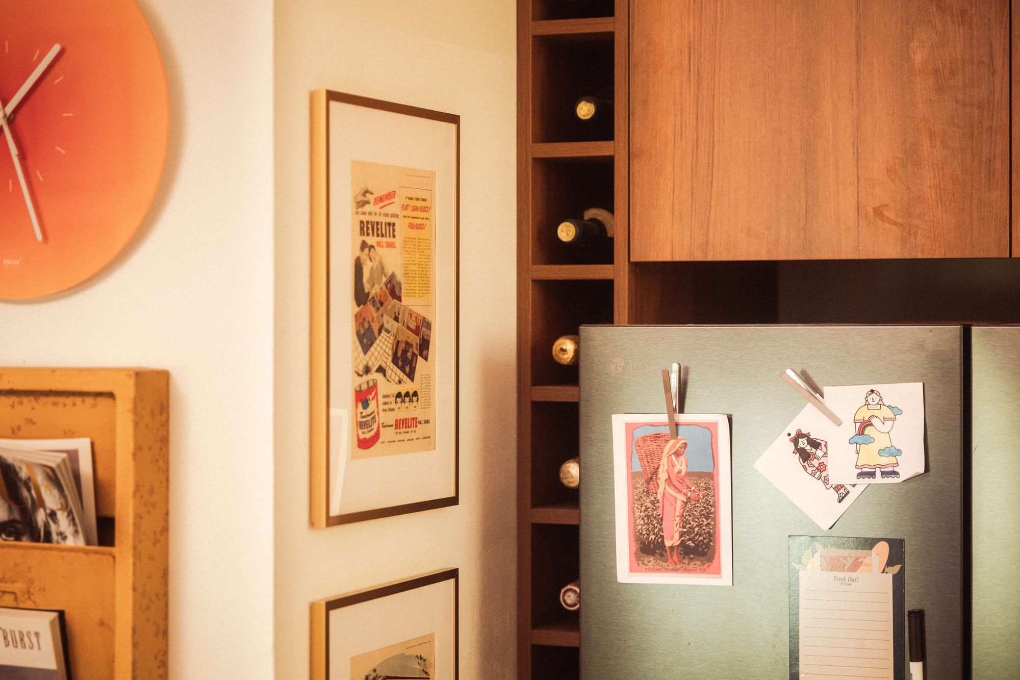 A kitchen corner detail with an orange wall clock, a framed vintage advertisement, warm timber cabinetry with integrated wine storage, and a stainless steel fridge door decorated with postcards and children's drawings clipped with pegs.