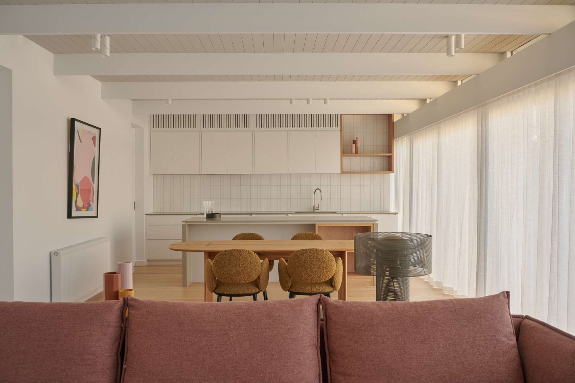 An open-plan living and kitchen space viewed from behind a pink sofa, with exposed timber ceiling beams, white cabinetry, an oval timber dining table with mustard bouclé chairs, and sheer floor-to-ceiling curtains.