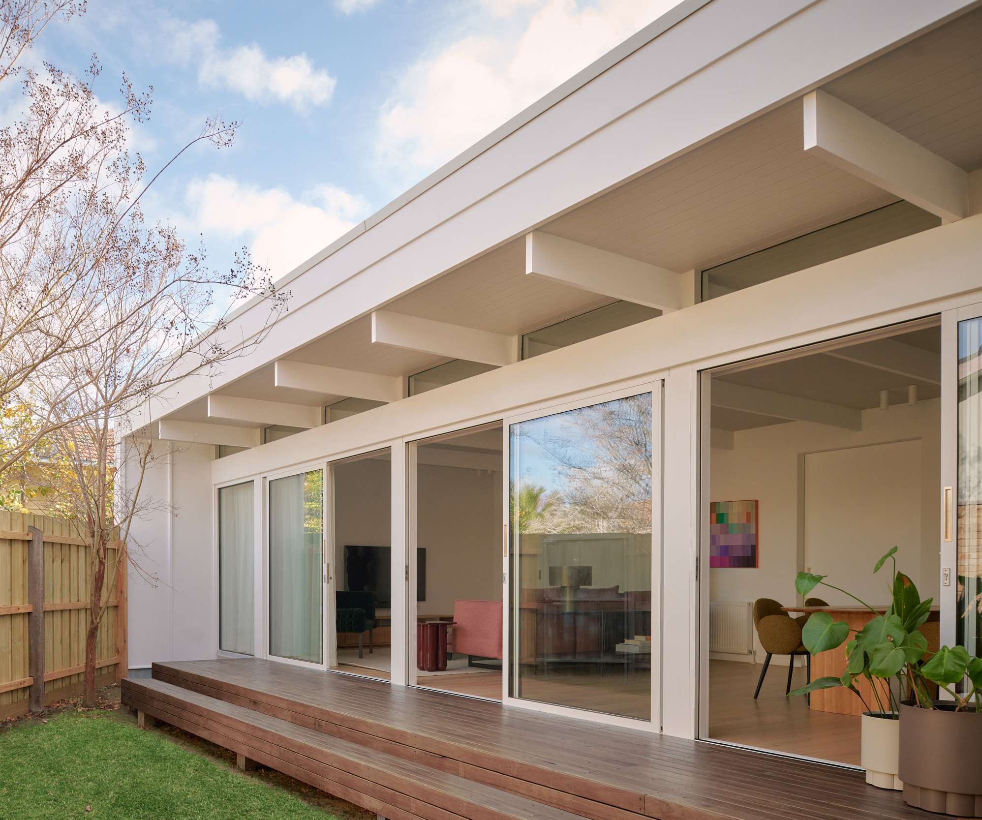 The rear facade of a mid-century inspired home with a wide overhanging roof, full-width sliding glass doors opening to a timber deck, green lawn, and mature trees visible against a blue sky.