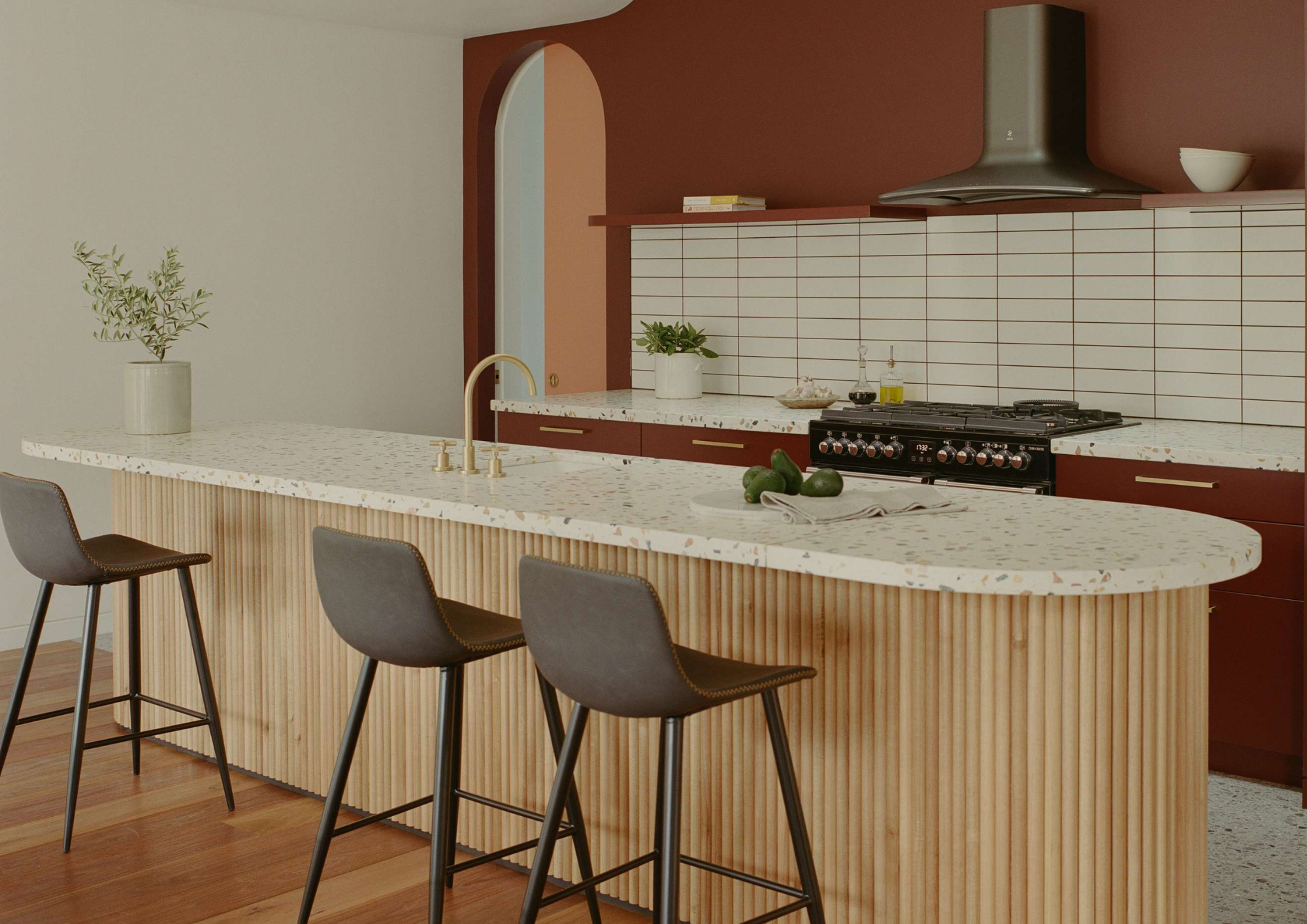 Kitchen with a terrazzo island, ribbed timber cabinetry and an ox blood coloured wall with an arched doorway in Northcote Hacienda by Hindley & Co