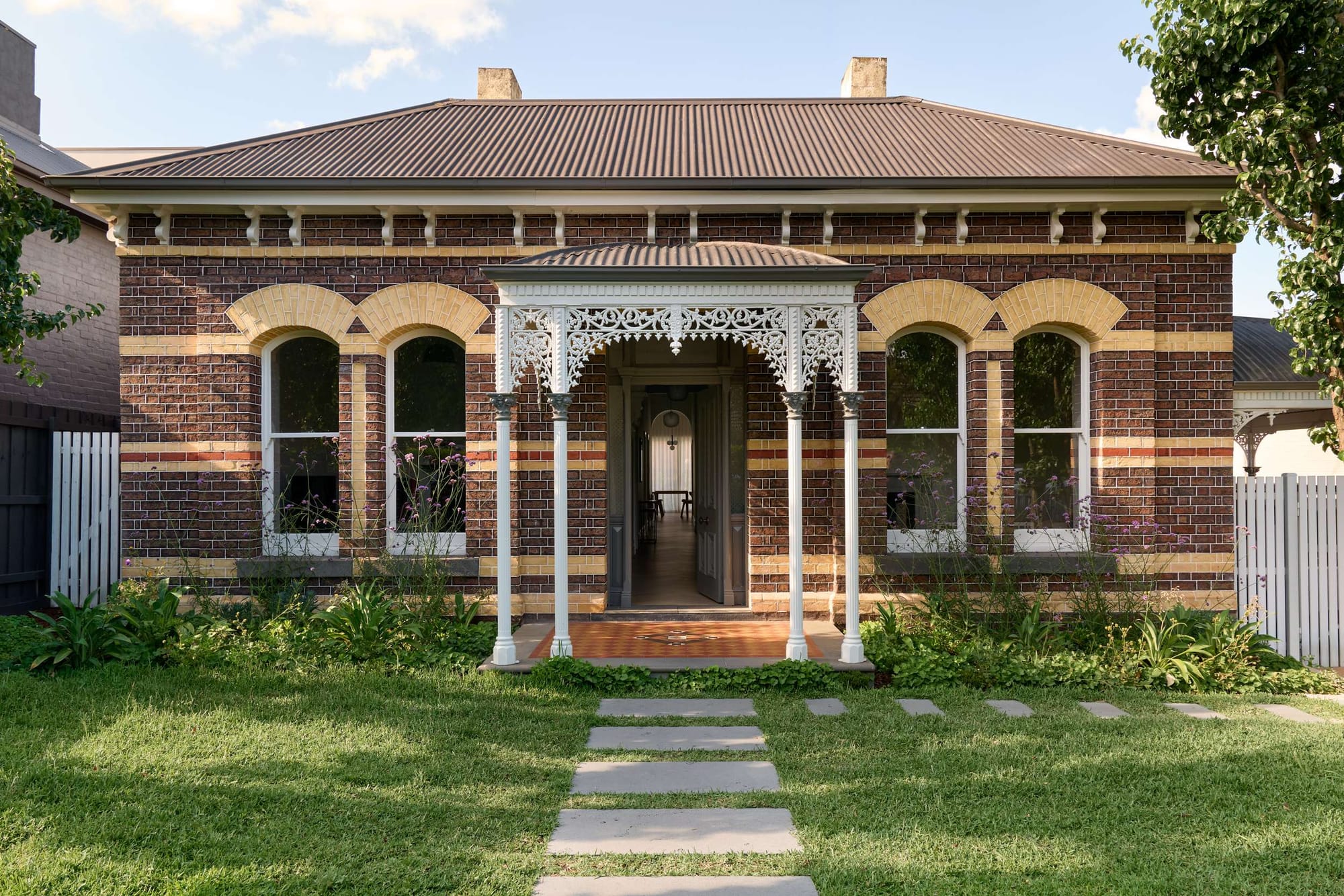 Polychrome brick Victorian villa with arched, tuck-pointed window surrounds and an ornate white iron lace entry portico, flanked by a landscaped garden and stepping-stone path.