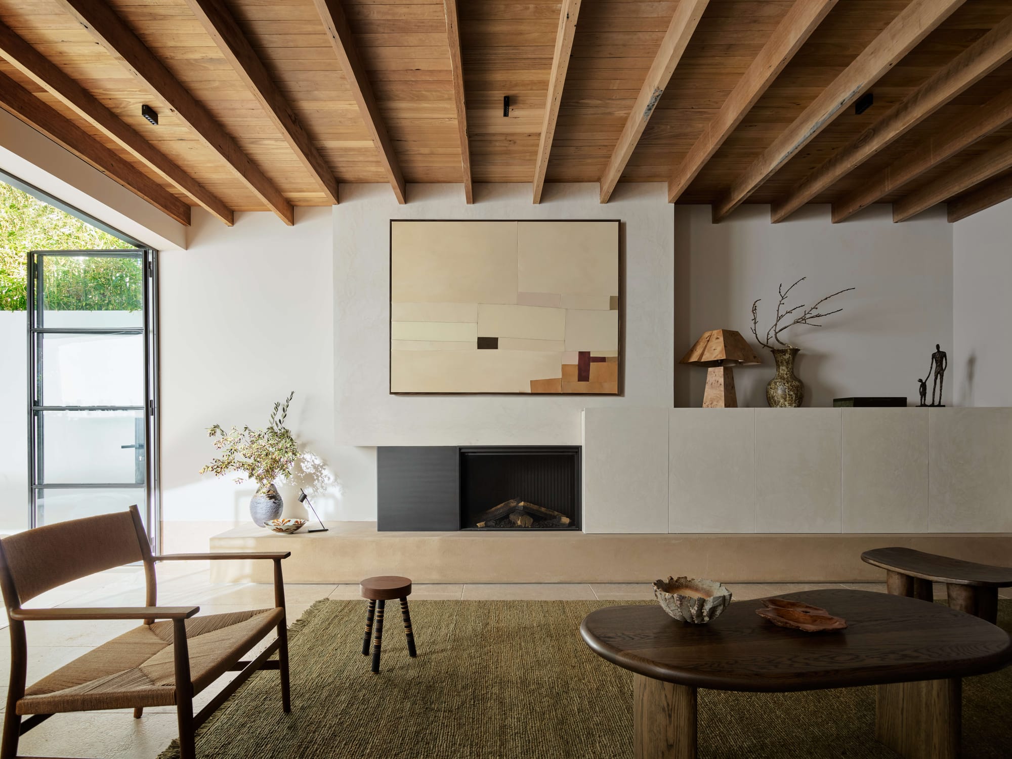Living room with a timber raked ceiling and exposed beams, a plaster fireplace surround with abstract artwork above, woven armchair and dark timber coffee table on a green rug.