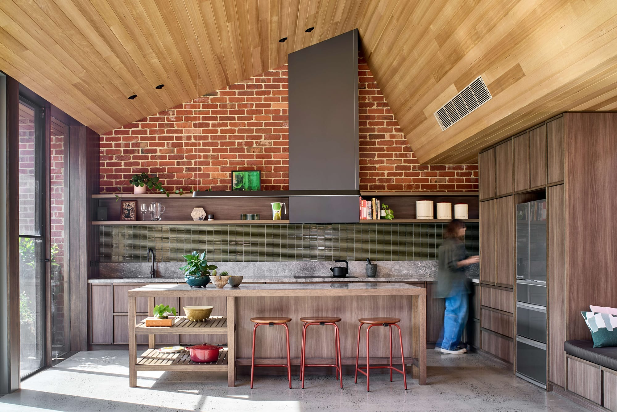 Vaulted timber ceiling kitchen with an exposed brick feature wall, green tiled splashback, and a timber island with red stools.