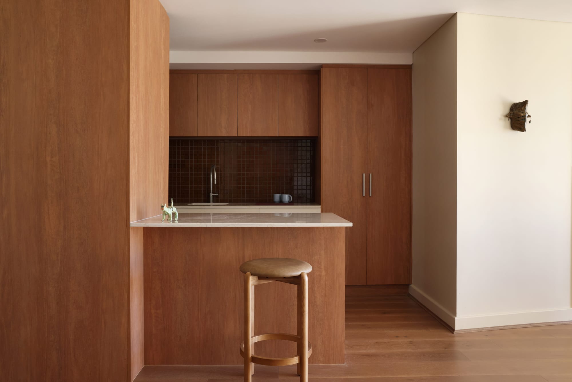 Timber panelled kitchen with a stone topped island, dark mosaic tile splashback, and a curved timber bar stool.