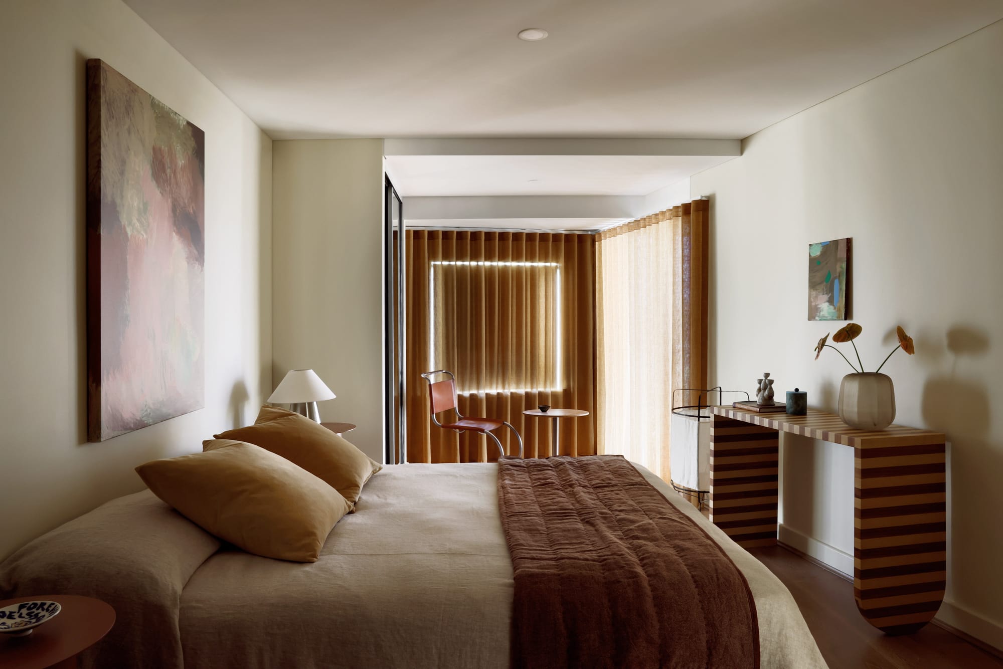 Bedroom with a mirrored wardrobe reflecting sheer amber curtains, a leather sling chair beside a small round table, abstract artwork above the bed and a striped bedside cabinet.