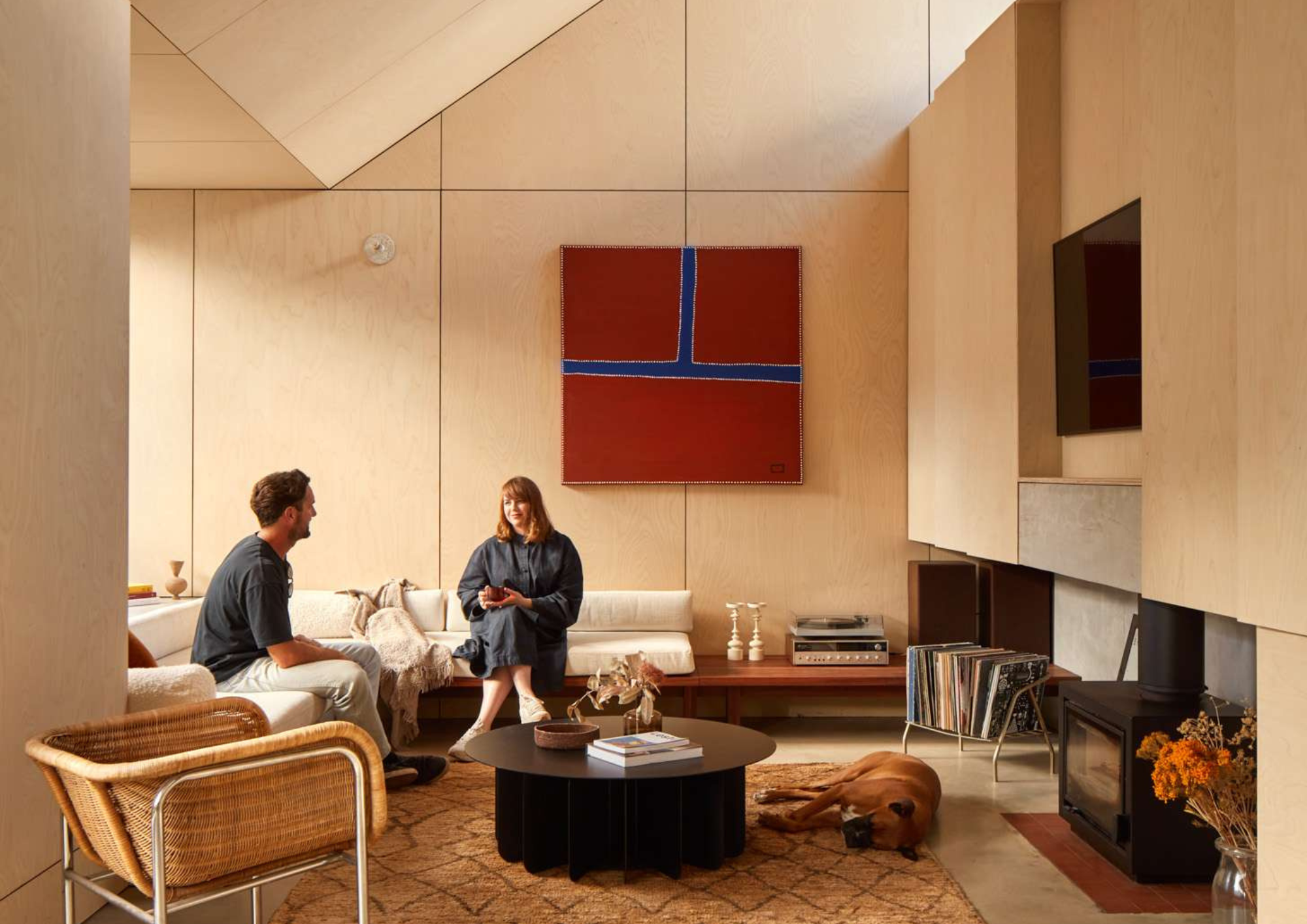 Living room with plywood lined walls, a wood fireplace, and two people seated on a bench sofa beneath framed artwork, with a dog resting nearby in Shadow House by Grotto Studio