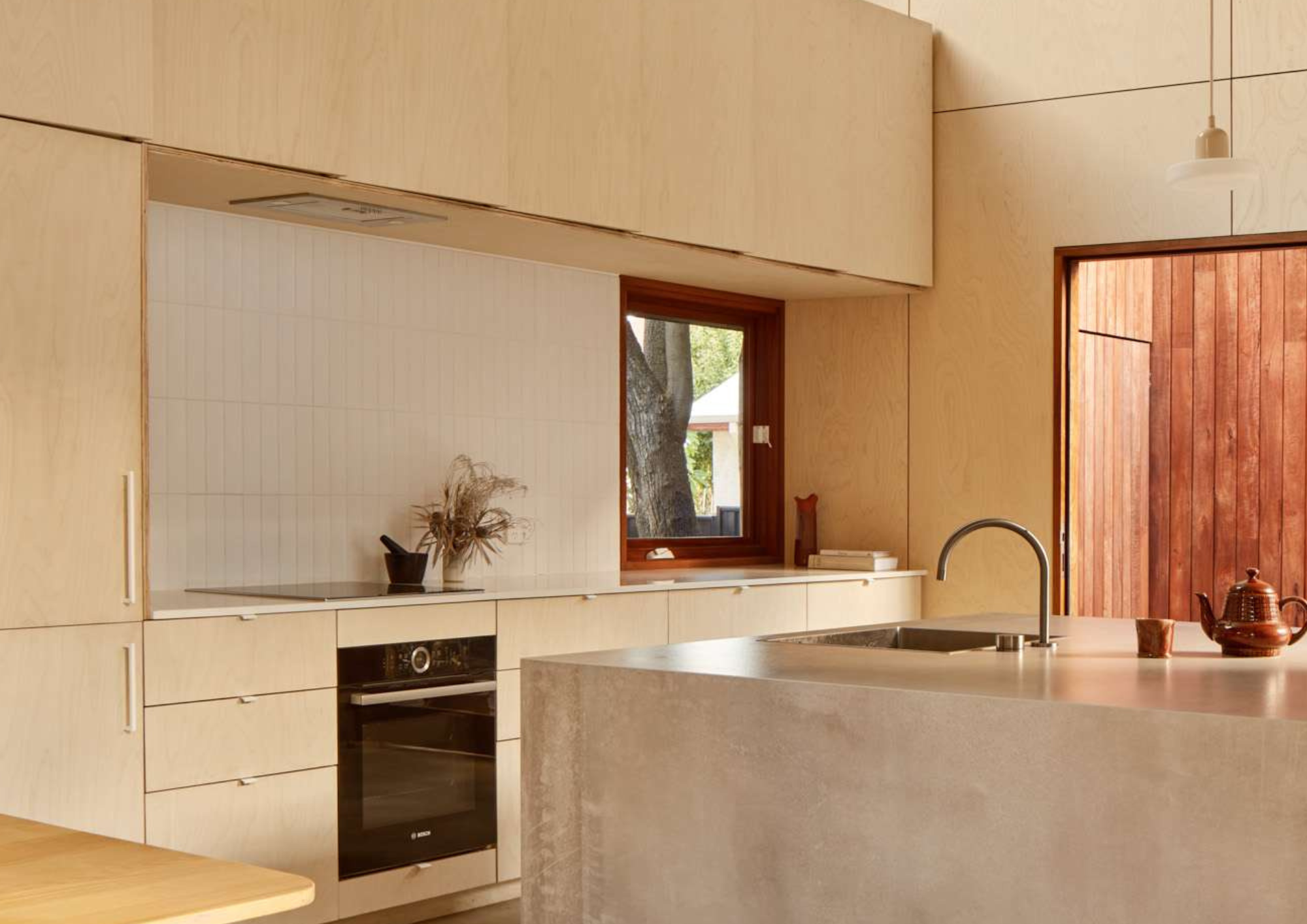Light timber plywood kitchen with white tiled splashback, built in oven and a warm red timber door opening to the garden in Shadow House by Grotto Studio