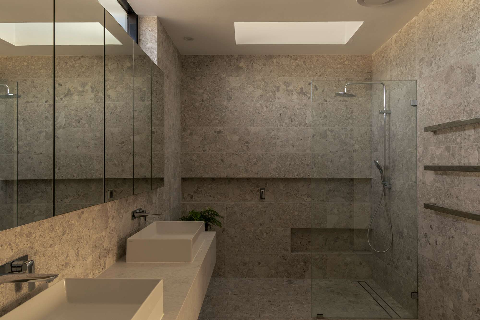 Stone-tiled bathroom with a frameless glass shower, mirrored cabinetry and twin vessel basins, by Keen Architecture.