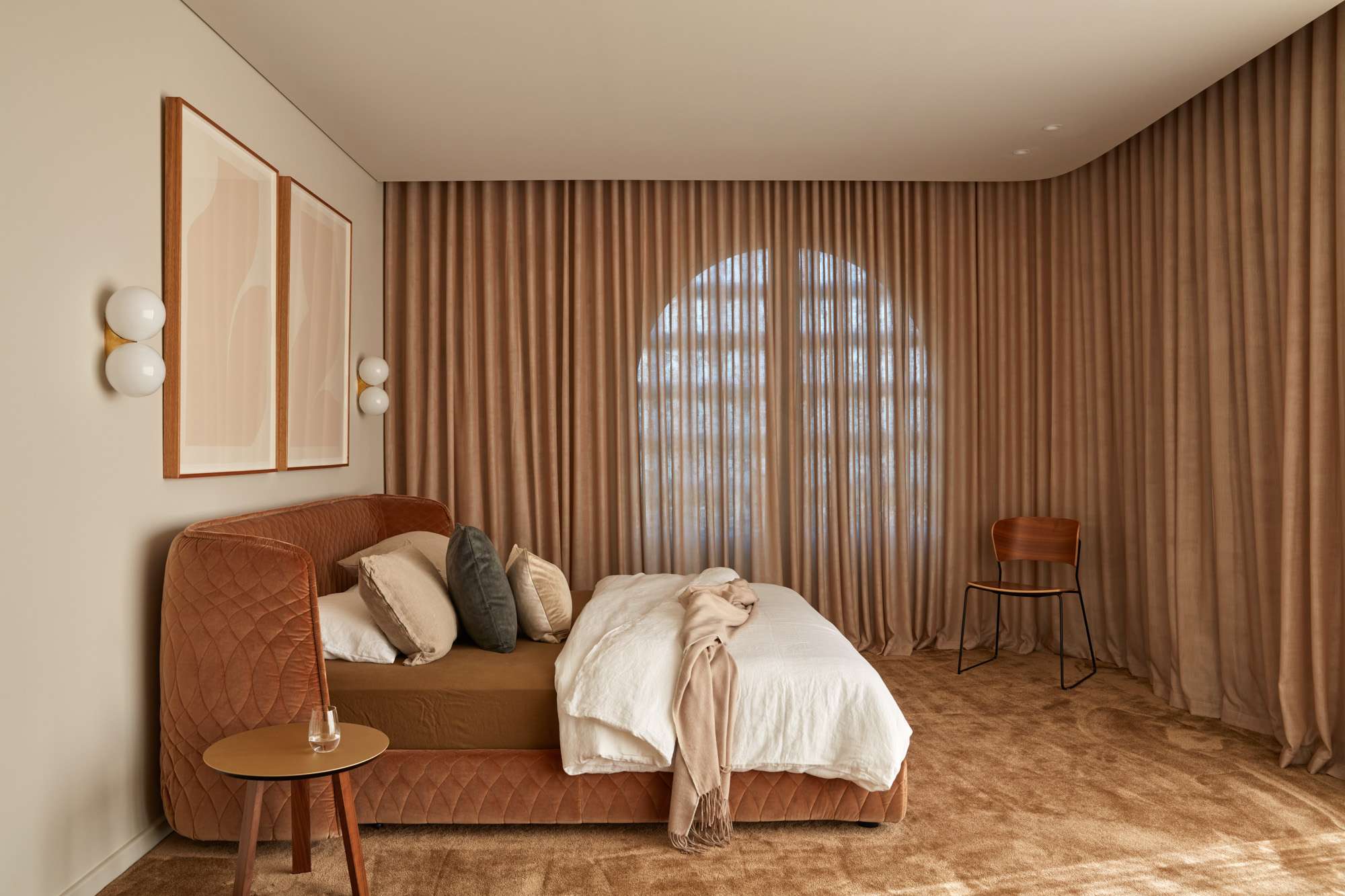 Bedroom with a curved quilted velvet bedhead in terracotta, layered cushions in charcoal and cream, arched window softened by floor to ceiling curtains, twin wall sconces beside framed artwork.
