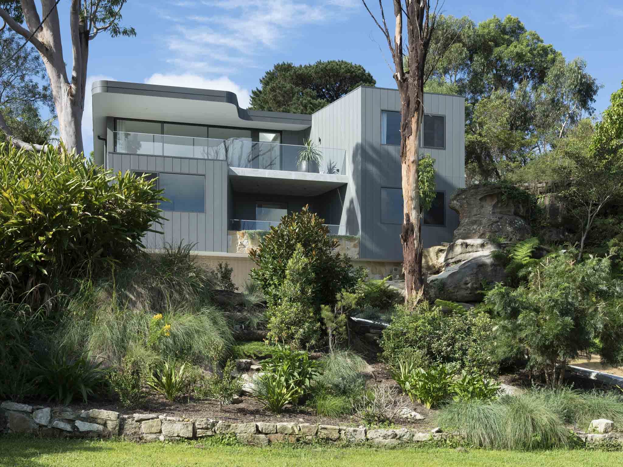 A side view of the grey clad multi-level home set into a sloped native garden, with sandstone boulders, tree ferns, and mature eucalyptus trees surrounding the glazed balconies and terraces.