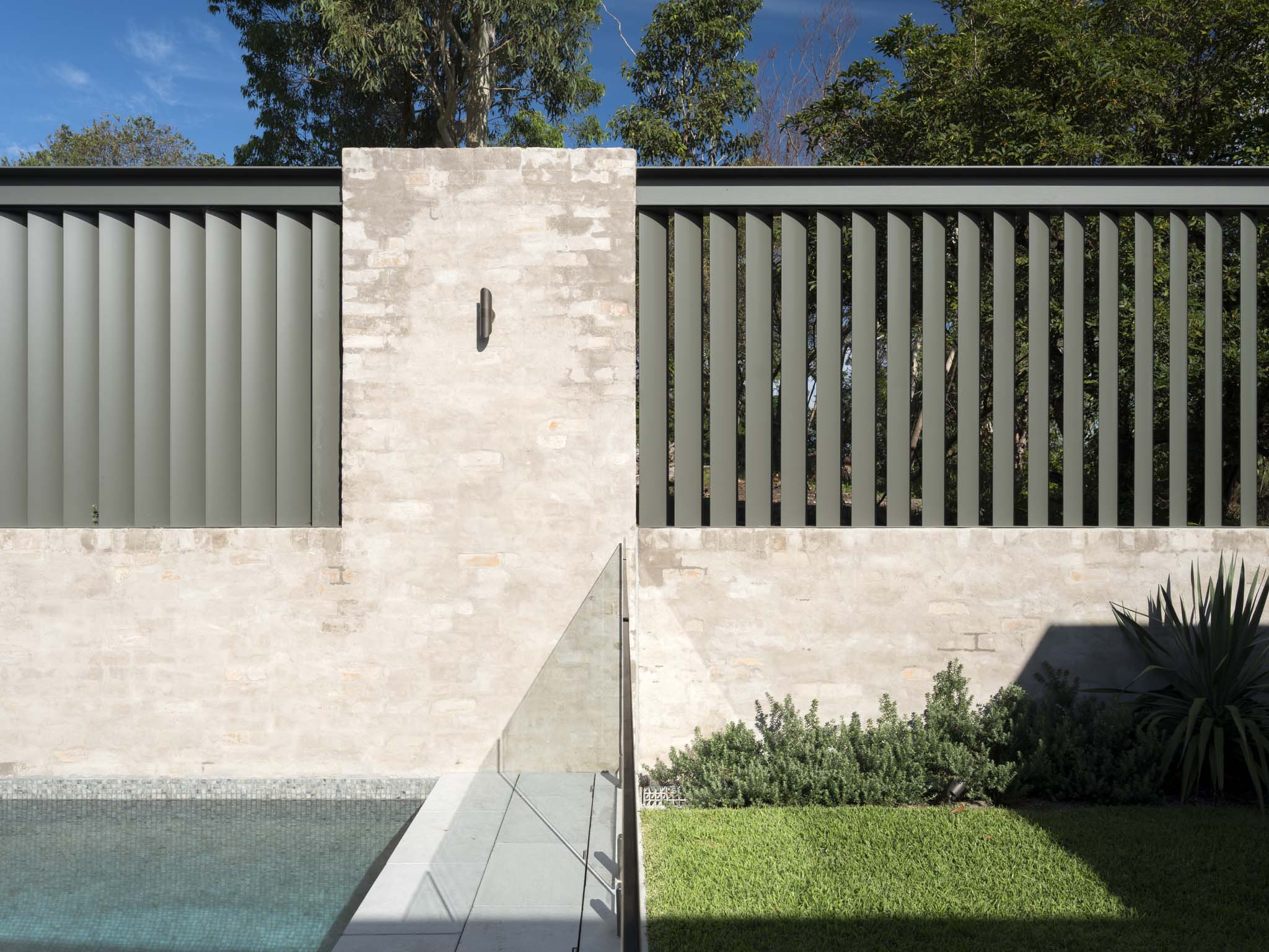 A pool-side boundary wall detail featuring a sandstone masonry pier flanked by dark grey vertical blade screening, with a glass pool fence, lawn, and native plantings in the foreground.