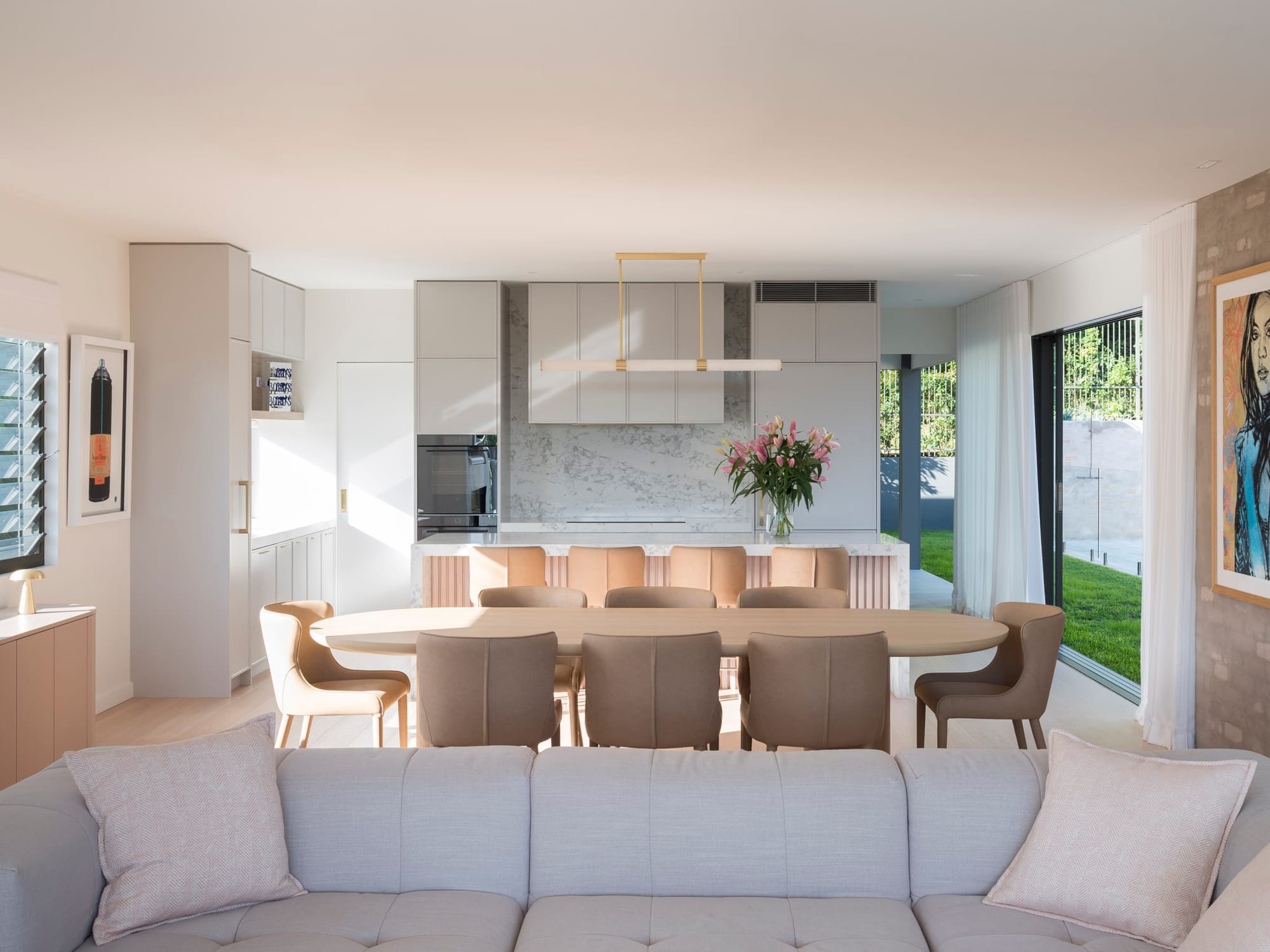 A bright open-plan kitchen and dining area with grey cabinetry, a marble splashback and island bench, an oval timber dining table with upholstered chairs, a brass pendant light, and sheer curtains framing a garden view.