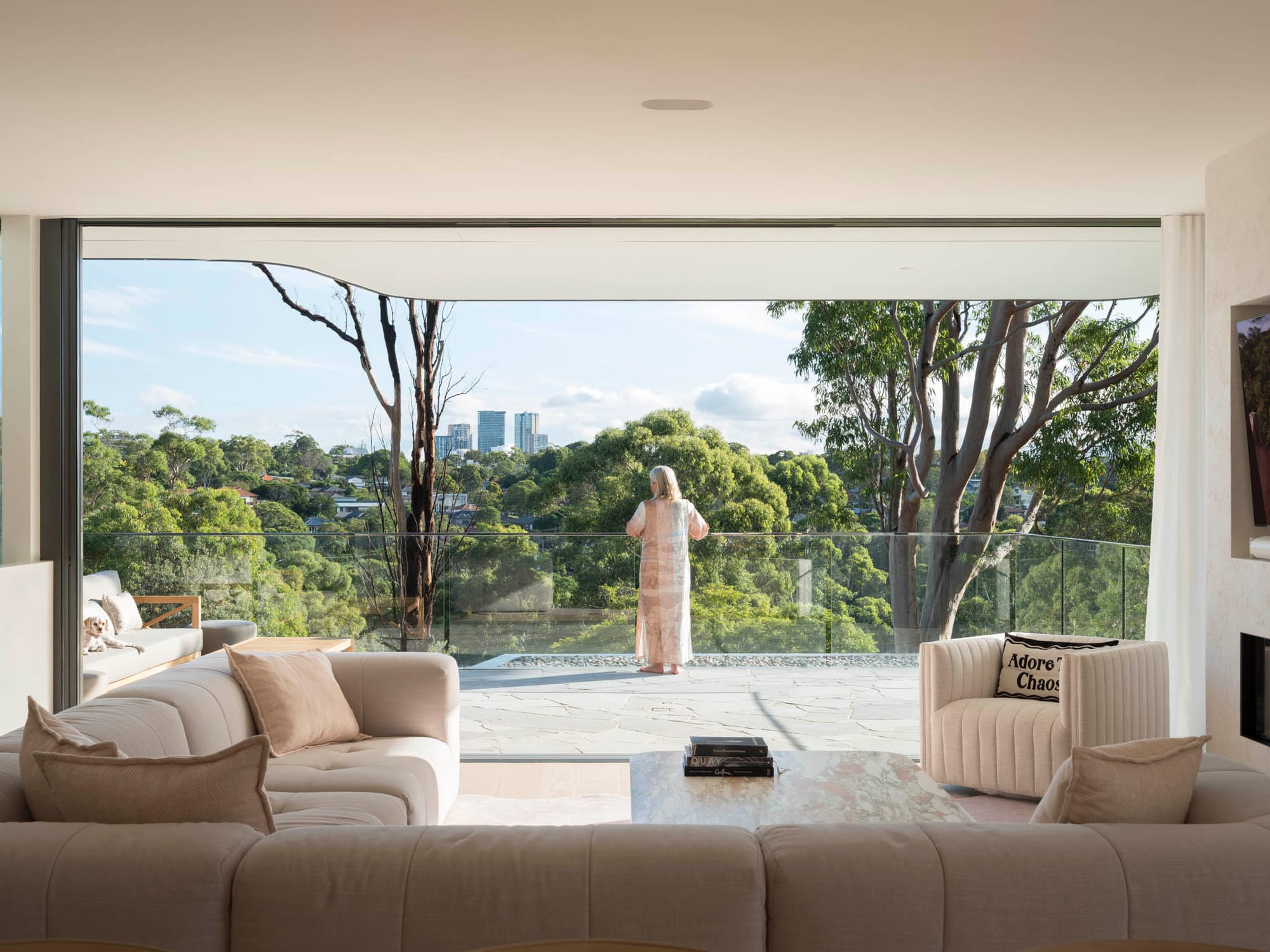 A light-filled living room opens fully to a cantilevered terrace through a wide frameless glass wall, with a woman standing at the glass balustrade looking out over a treetop and city skyline view.
