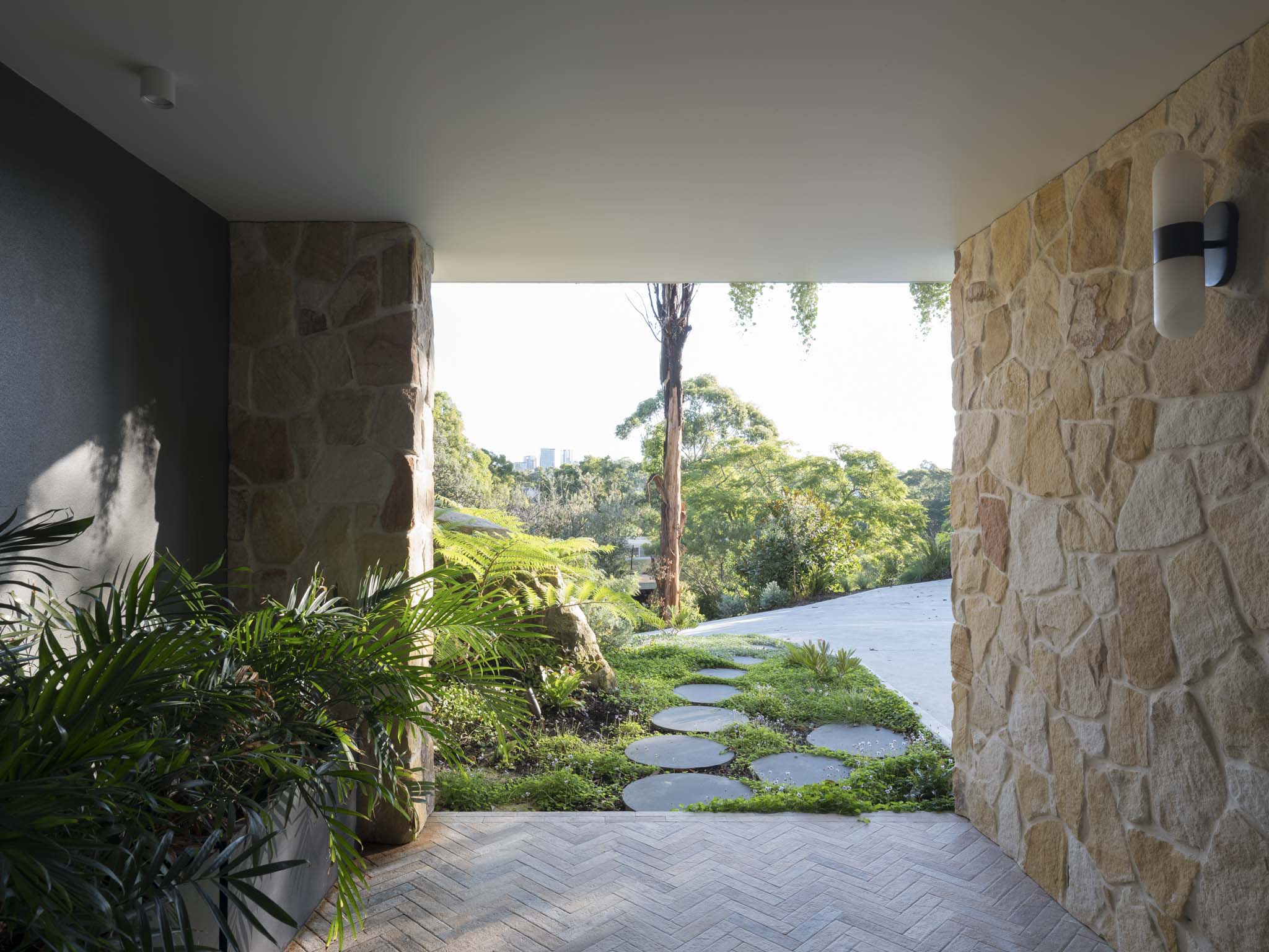 A covered entry portico with sandstone feature walls, herringbone brick paving, and circular stepping stones leading through a lush fern garden toward a framed view of eucalyptus trees and the city skyline beyond.
