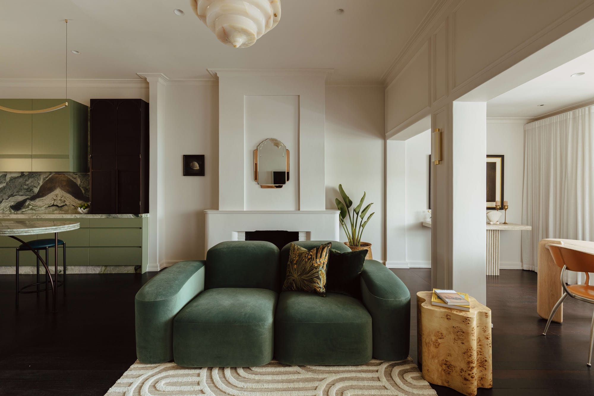Living room with a rounded green velvet sofa, ornate fireplace mantel, sculptural mirror, and sage green kitchen visible beyond.