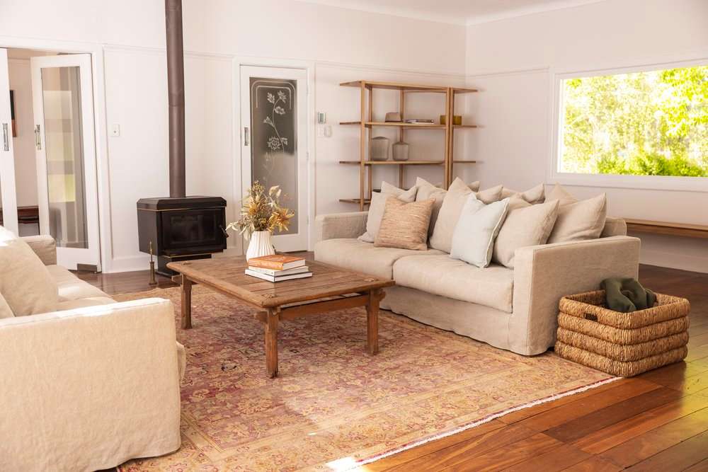 Living room in a country style home with a freestanding wood heater, linen slipcovered sofas, a vintage timber coffee table on a faded rug and an open timber shelving unit.