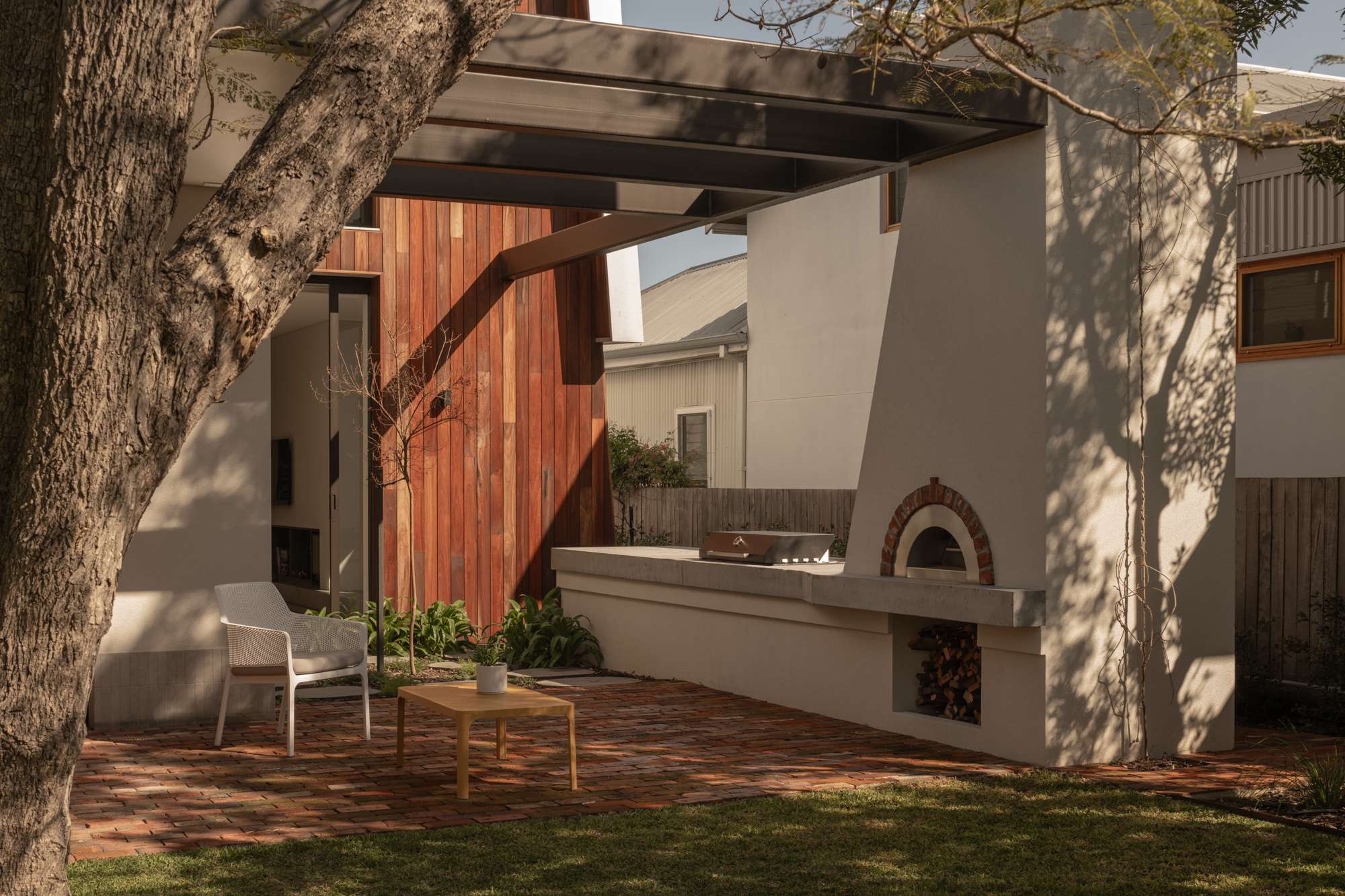 Outdoor entertaining area with a built in pizza oven, concrete benchtop, and steel awning, framed by a red timber clad wall and mature garden trees.