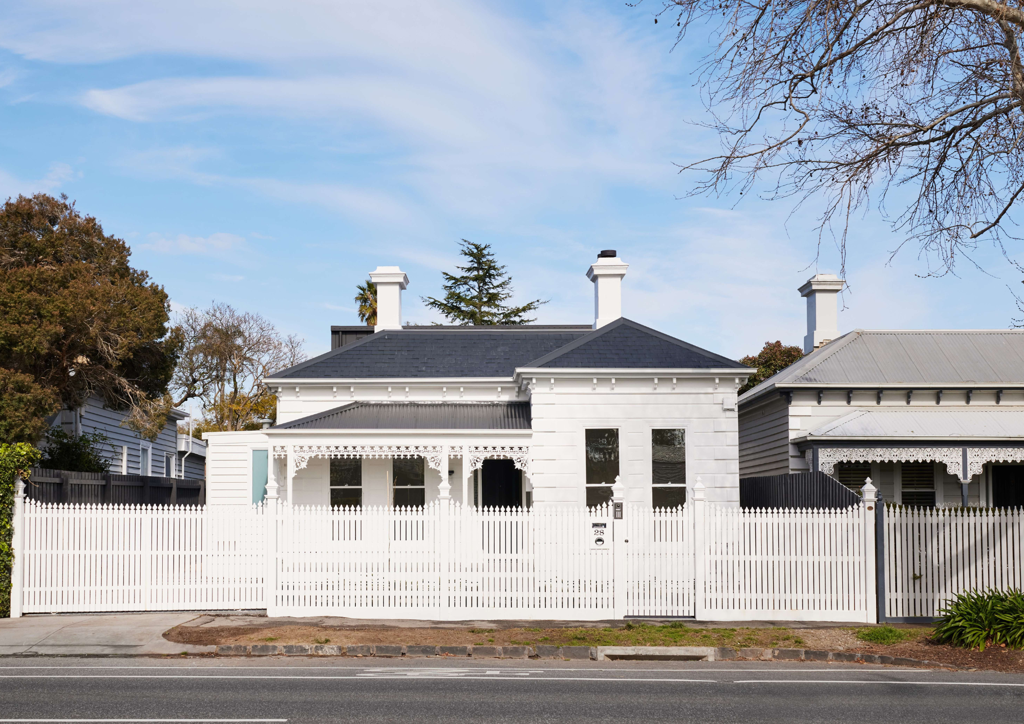 Daytime street view of a restored white Victorian villa with decorative eaves, twin chimneys and a scalloped picket fence.