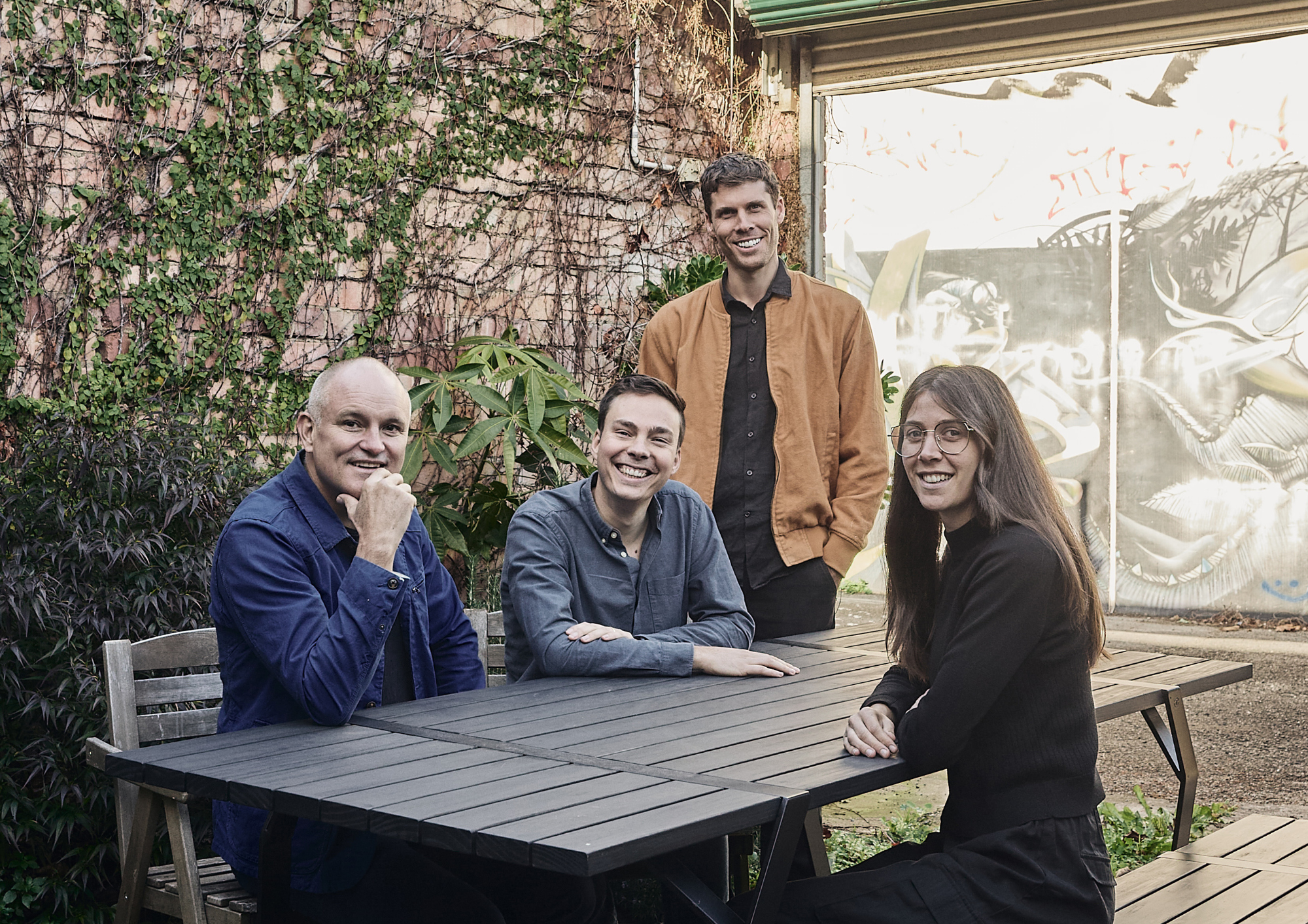 Four members of the MRTN Architects team around a picnic table, with an ivy-covered brick wall and graffiti mural behind them.