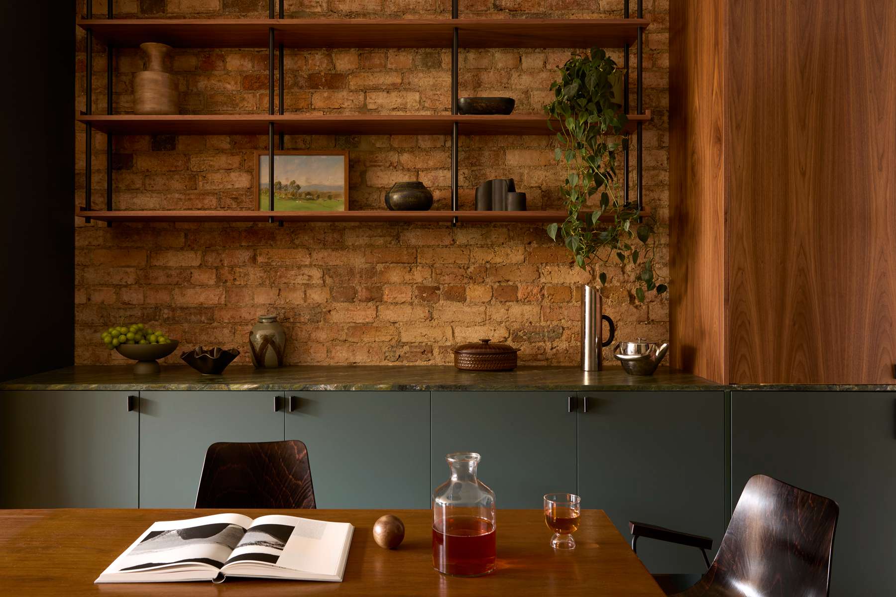 Exposed brick wall with open timber shelving, dark green stone benchtop, and a walnut dining table set with a book and carafe.