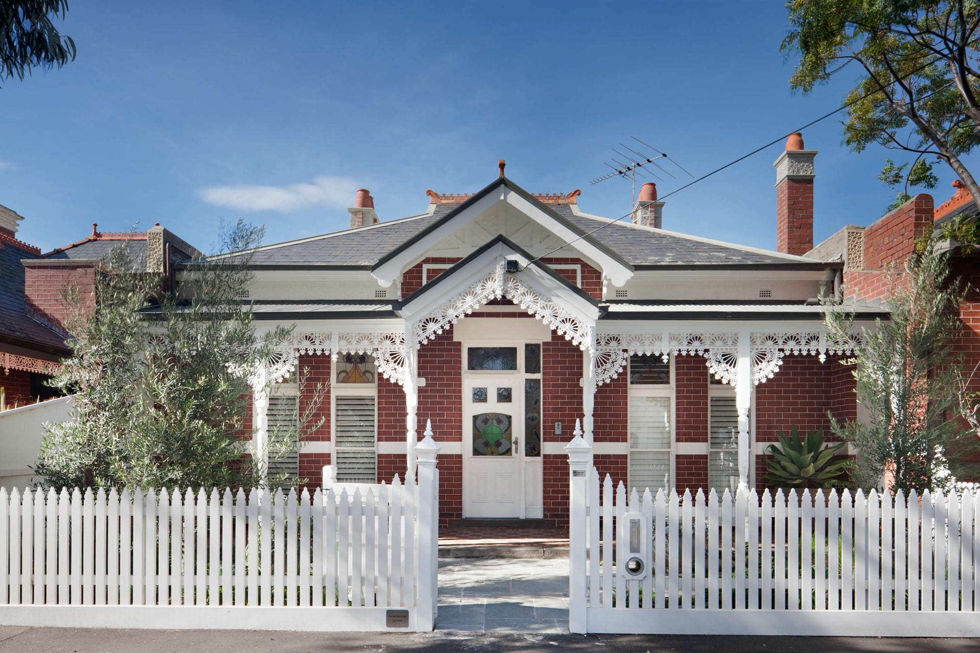 Red brick Victorian villa with intricate white lacework veranda detailing, a stained-glass front door and established garden plantings behind a picket fence.