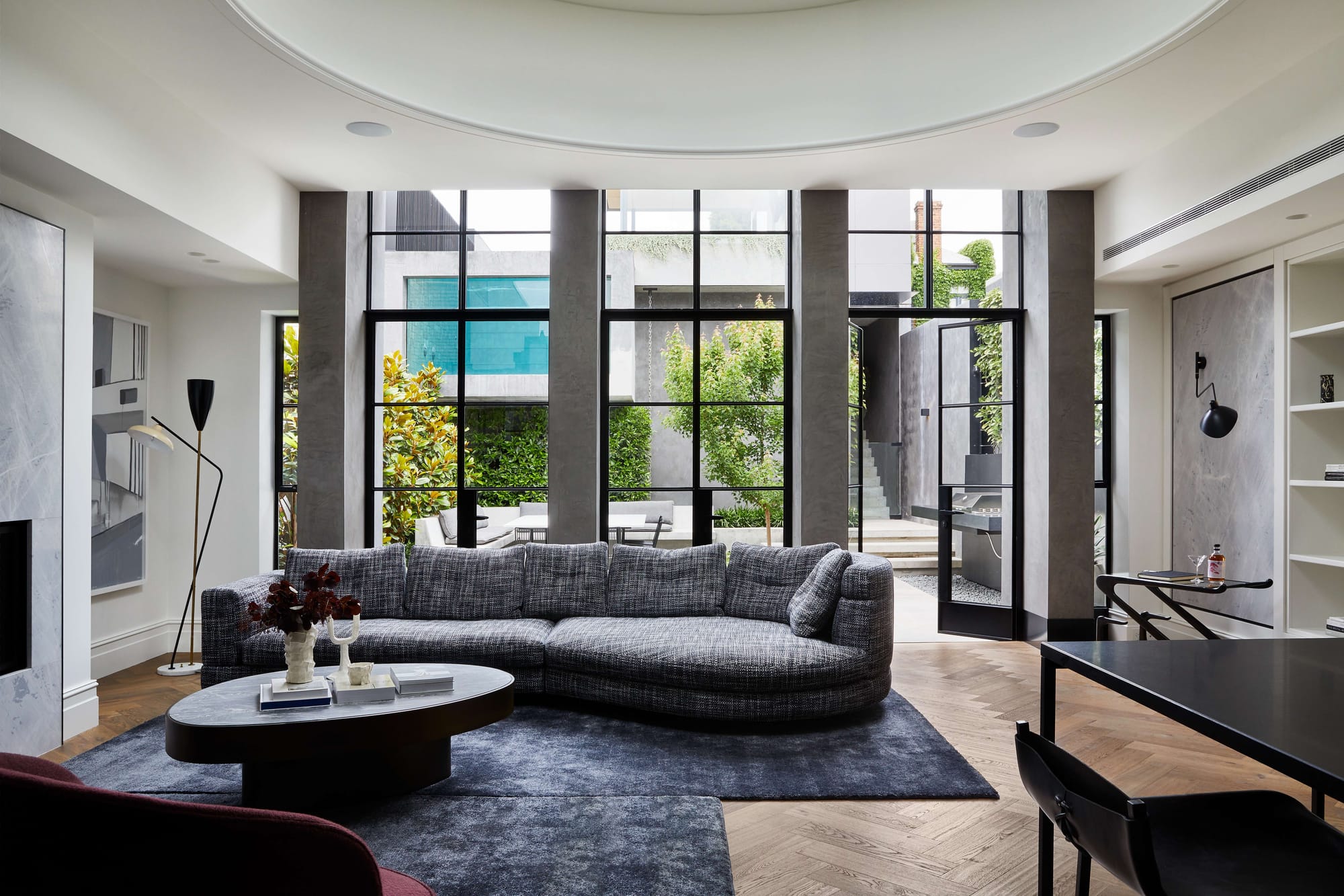 Living room with a curved grey tweed sofa, black oval coffee table and herringbone floors, framed by tall black steel windows opening onto a courtyard with plants and stairs.