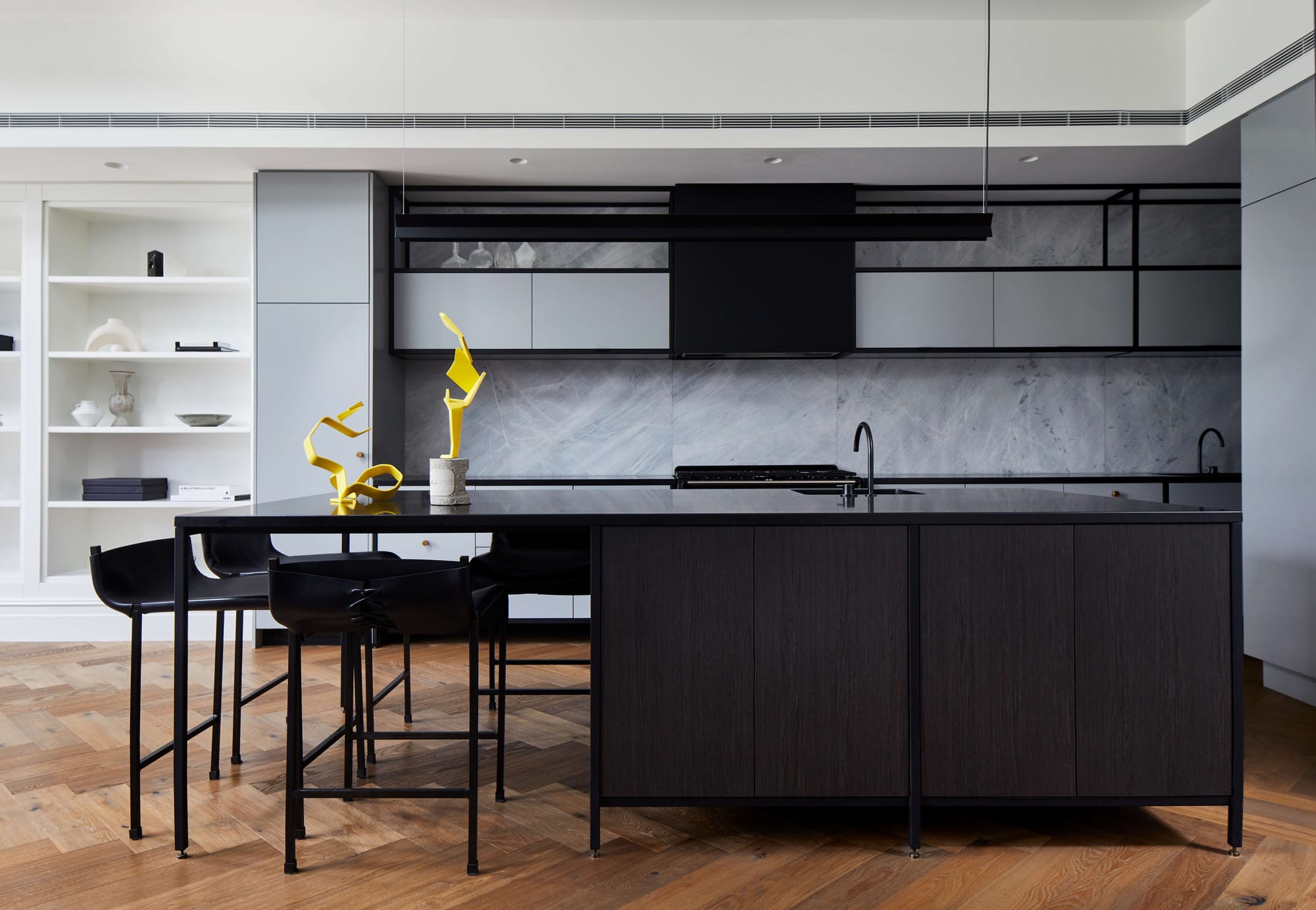 Kitchen with a dark timber island, black stone benchtop, grey marble splashback and black bar stools, styled with a pair of yellow sculptural objects.