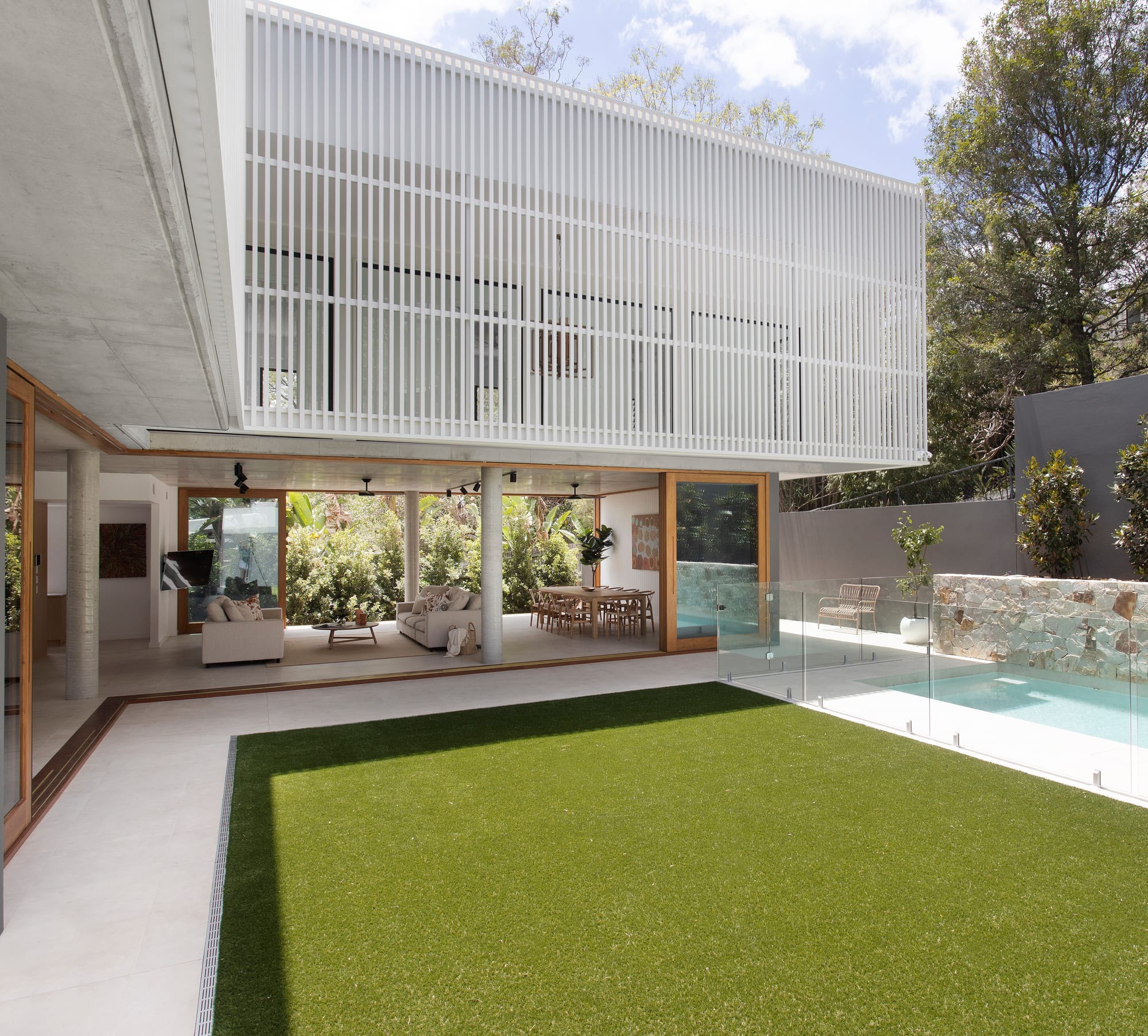 Modern home with white vertical screening overlooking a courtyard, lawn and swimming pool beside open-plan living spaces.