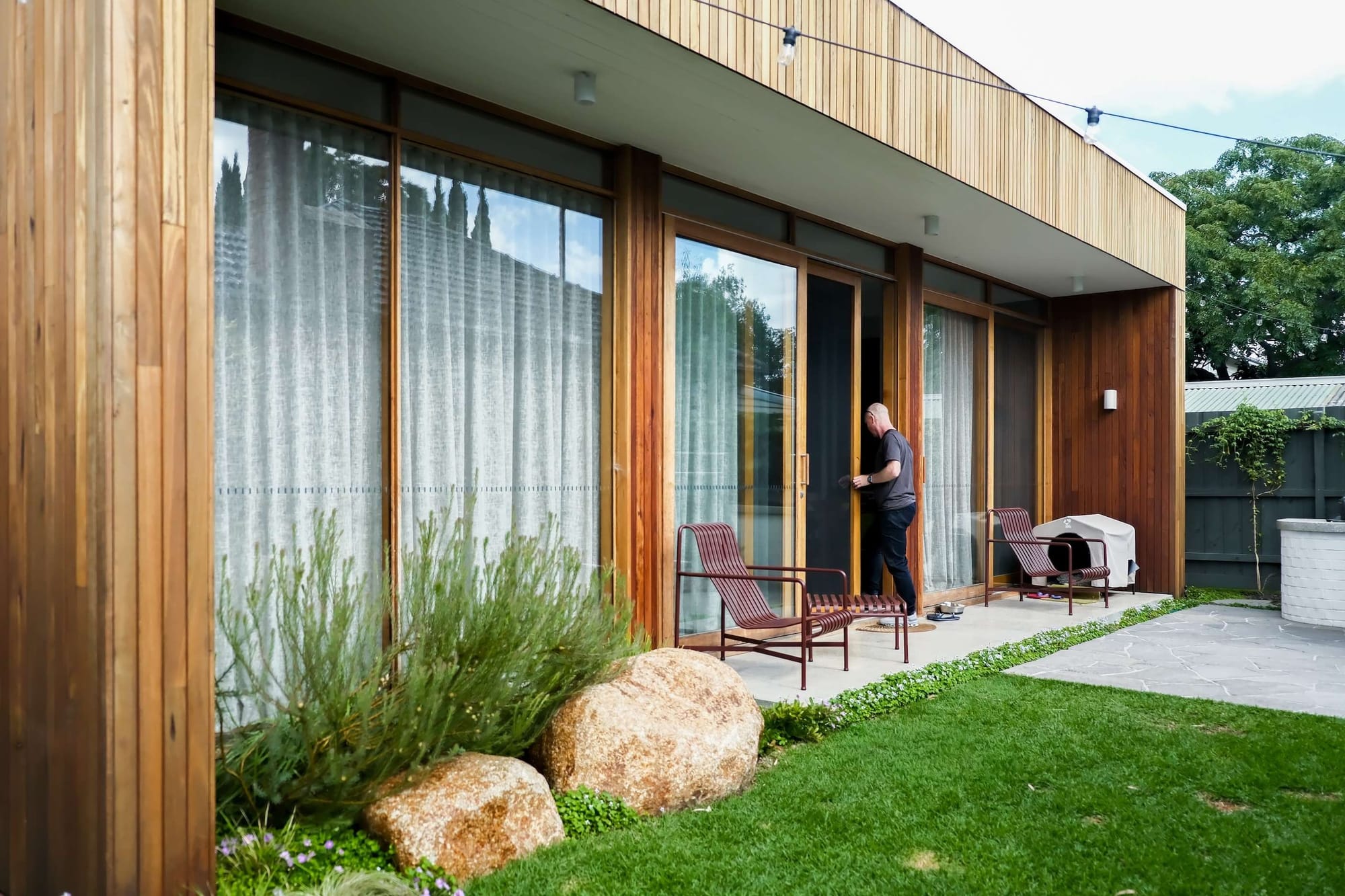 Timber clad home exterior with floor to ceiling glass doors, sheer curtains, a paved courtyard, garden beds with large rocks, and outdoor seating.