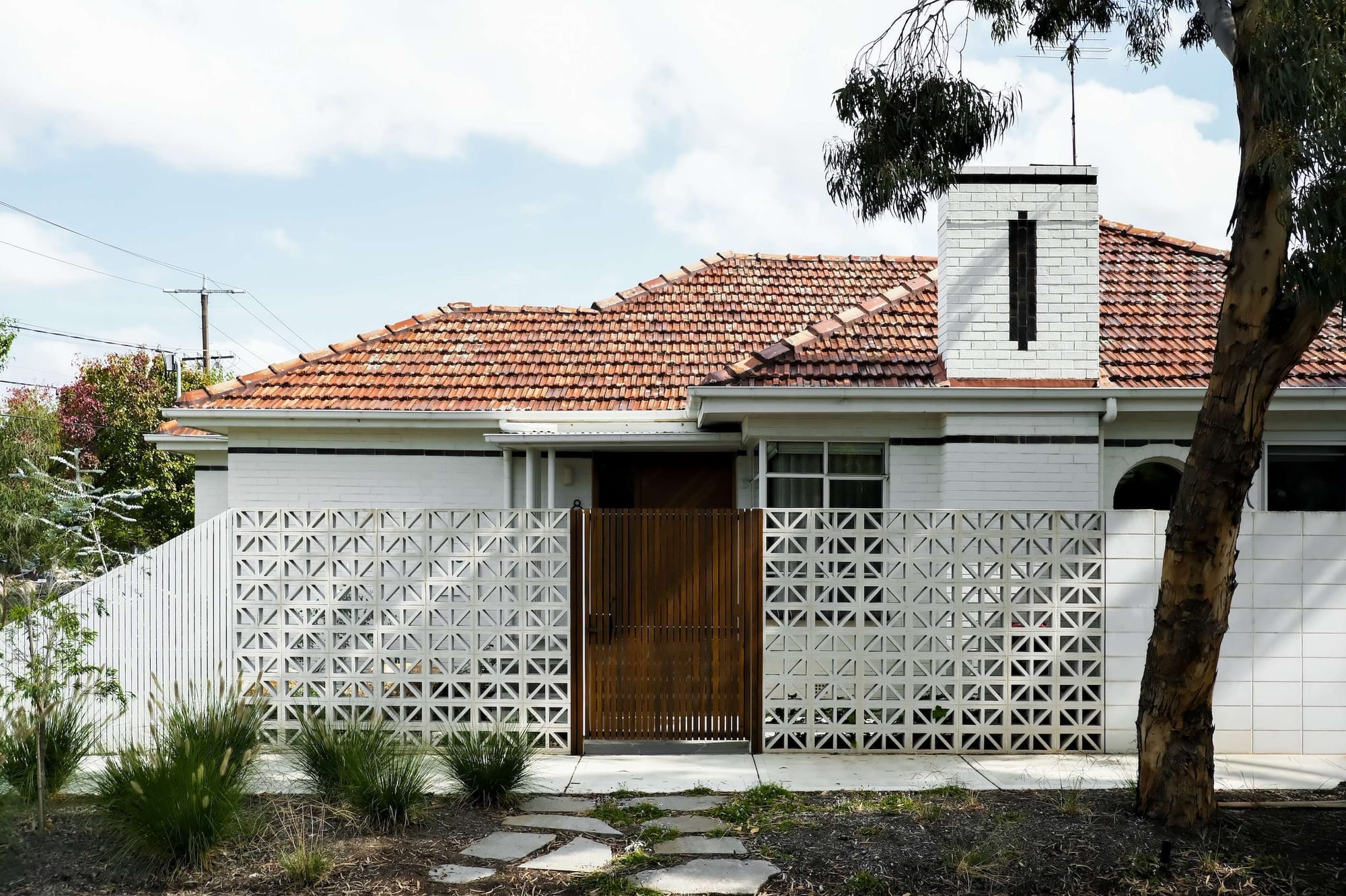 Renovated brick heritage home with a terracotta tiled roof, white chimney, decorative breeze block fencing and a timber slat gate, set among native grasses and a gum tree.