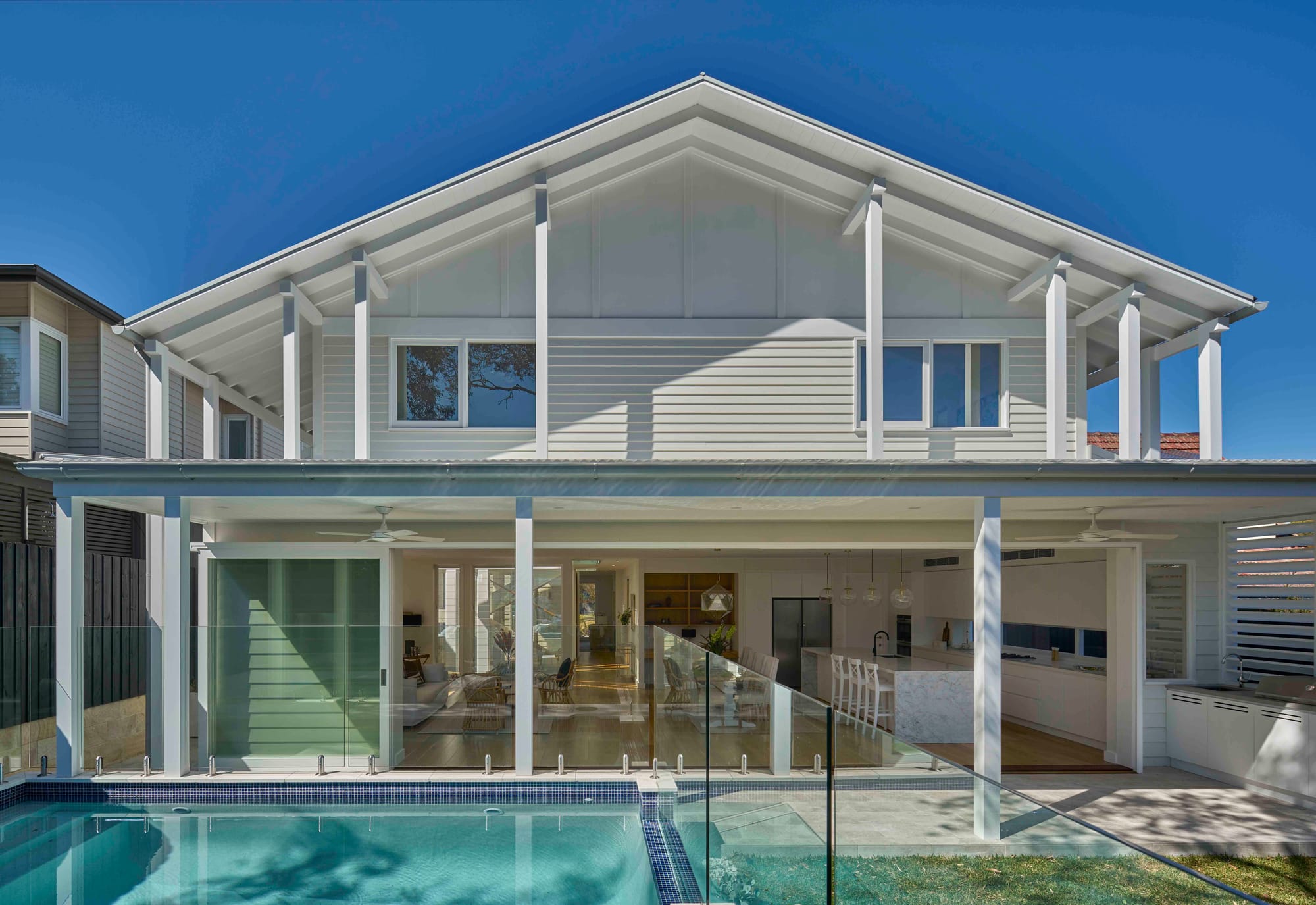 Rear view of a white weatherboard home with a gabled roof and exposed timber rafters, an open plan living and kitchen area behind glass sliding doors, and a tiled pool with glass balustrading in the foreground.