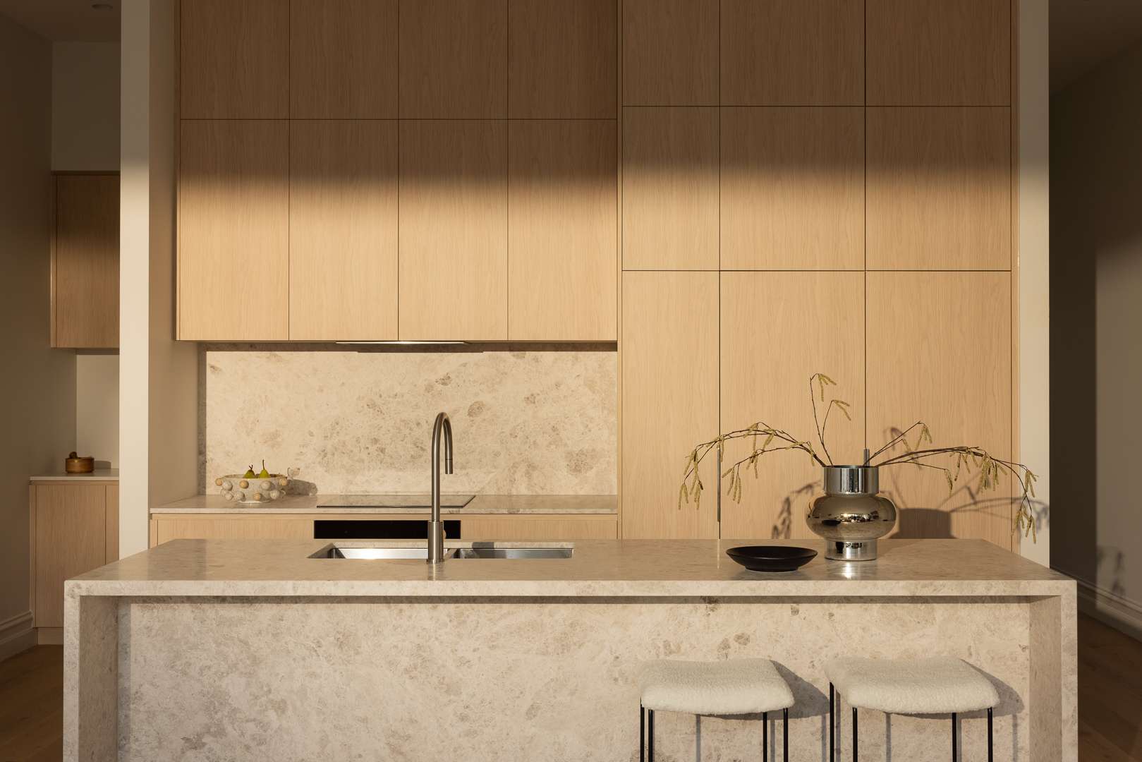 Contemporary kitchen with pale timber cabinetry, a stone island bench, integrated sink and warm afternoon light across the joinery.
