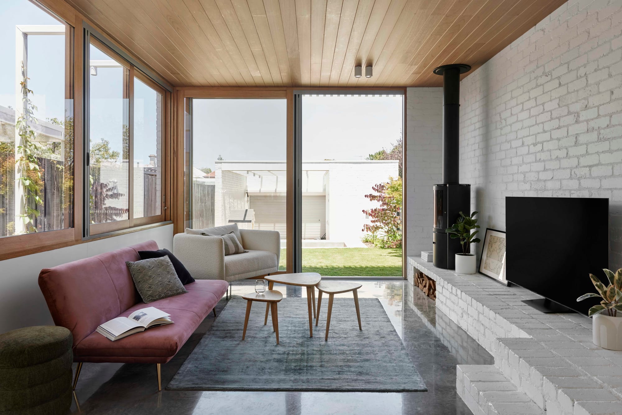 Compact living room in Rosie by Eckersley Architects with a timber ceiling, white brick fireplace, pink sofa and glazing to the garden.