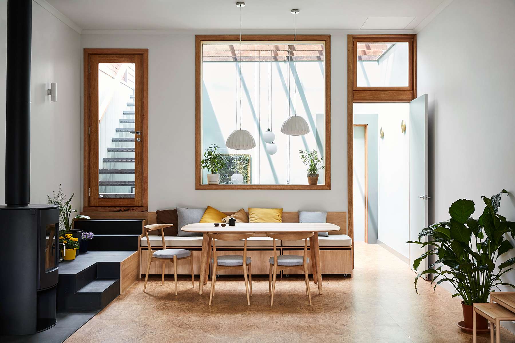 Light-filled dining space at Atop A Shop by Sustainable Homes Melbourne with a built-in timber banquette, pendant lights and black wood heater.