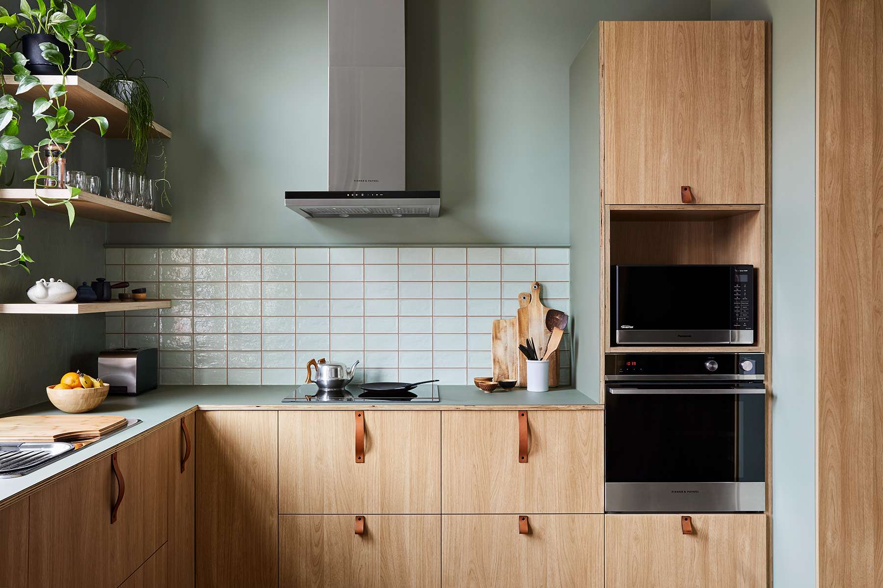 Compact green kitchen with timber cabinetry, open shelving, tiled splashback and integrated black appliances.