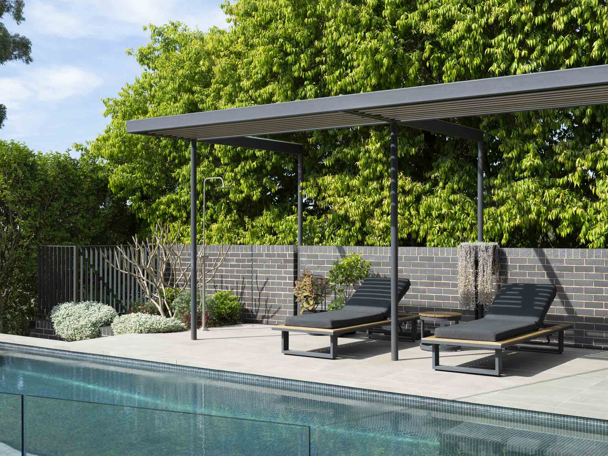 A timber-slatted poolside pergola shading two charcoal sun loungers and a side table, with an outdoor shower against a brick boundary wall and dense greenery behind.