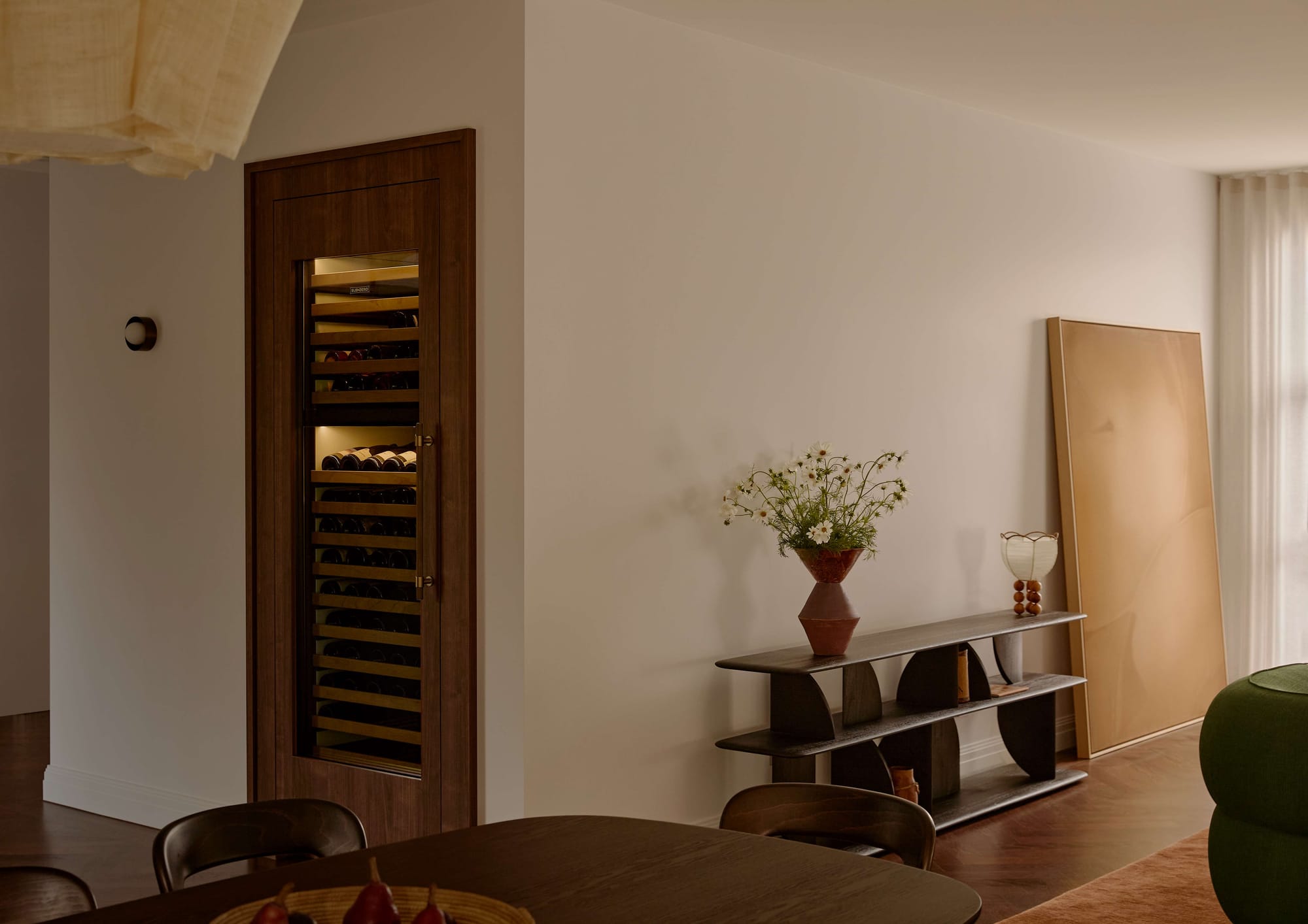 Built-in timber wine cabinet beside a black console table with sculptural legs, styled with a terracotta vase of white flowers and a leaning gold framed mirror.