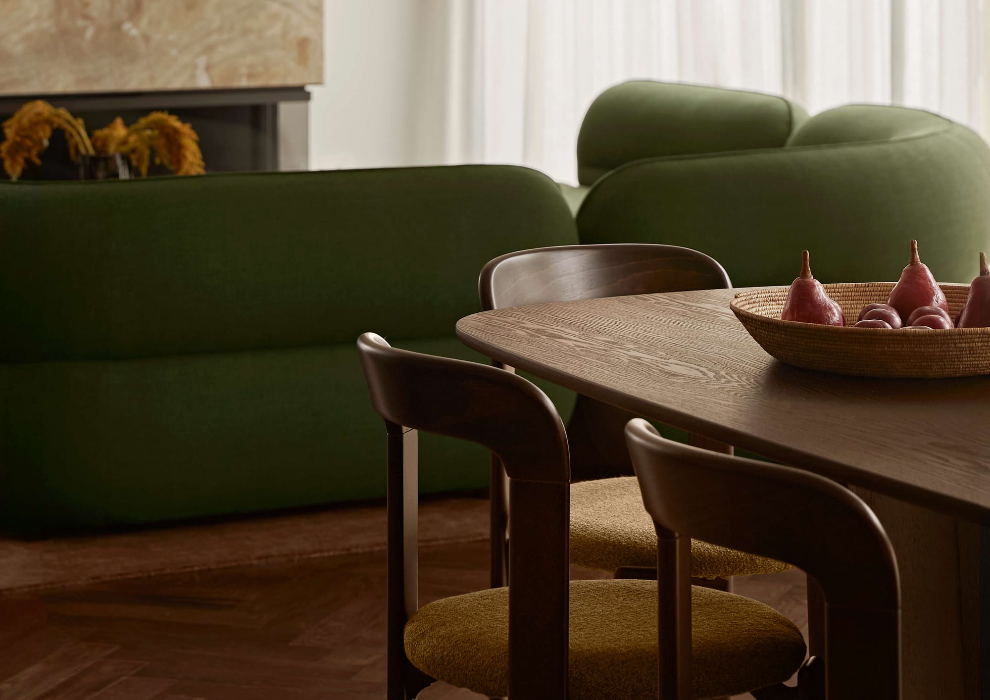 Close up of a dark timber dining table with curved wooden chairs in olive boucle upholstery, a woven bowl of pears, and a green curved sofa in the background.