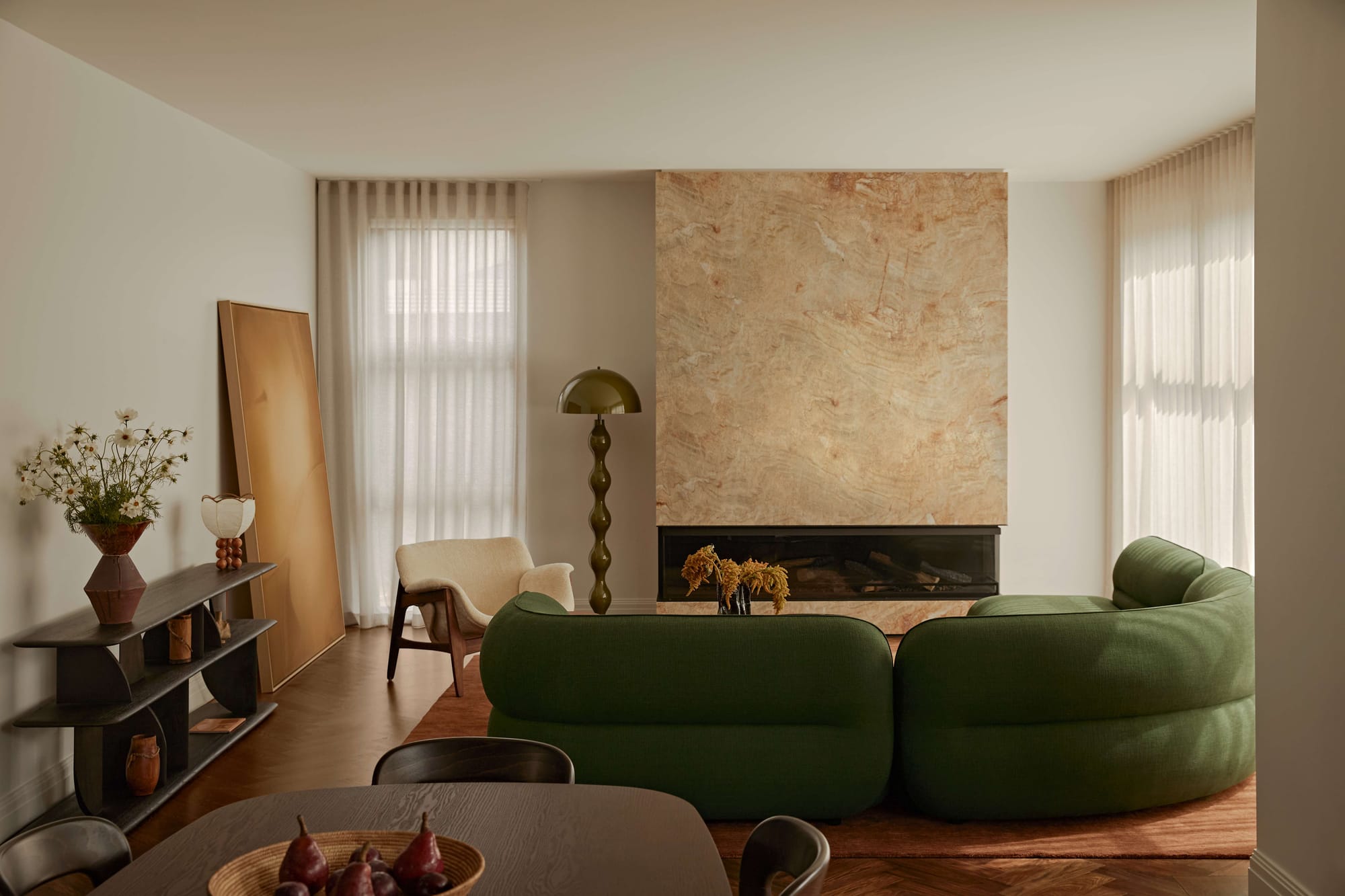 Living room with an olive green curved sofa, warm marble fireplace surround, cream armchair and sheer curtains, styled with dried florals and a sculptural floor lamp.