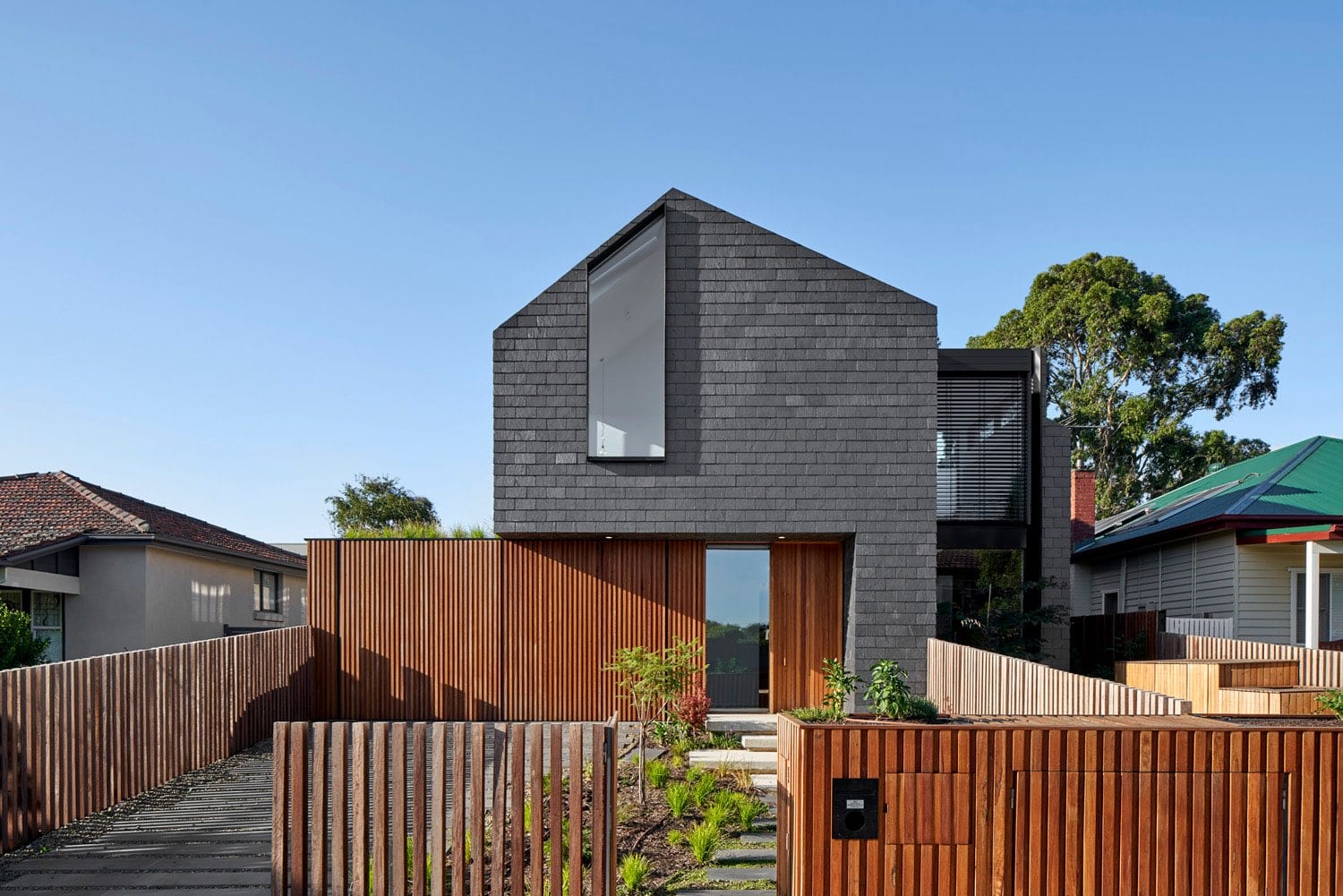 A grey brick gable-roofed house with a timber-slat fence and entry screen, a tall angled window at its peak, and a dark glazed upper extension behind.