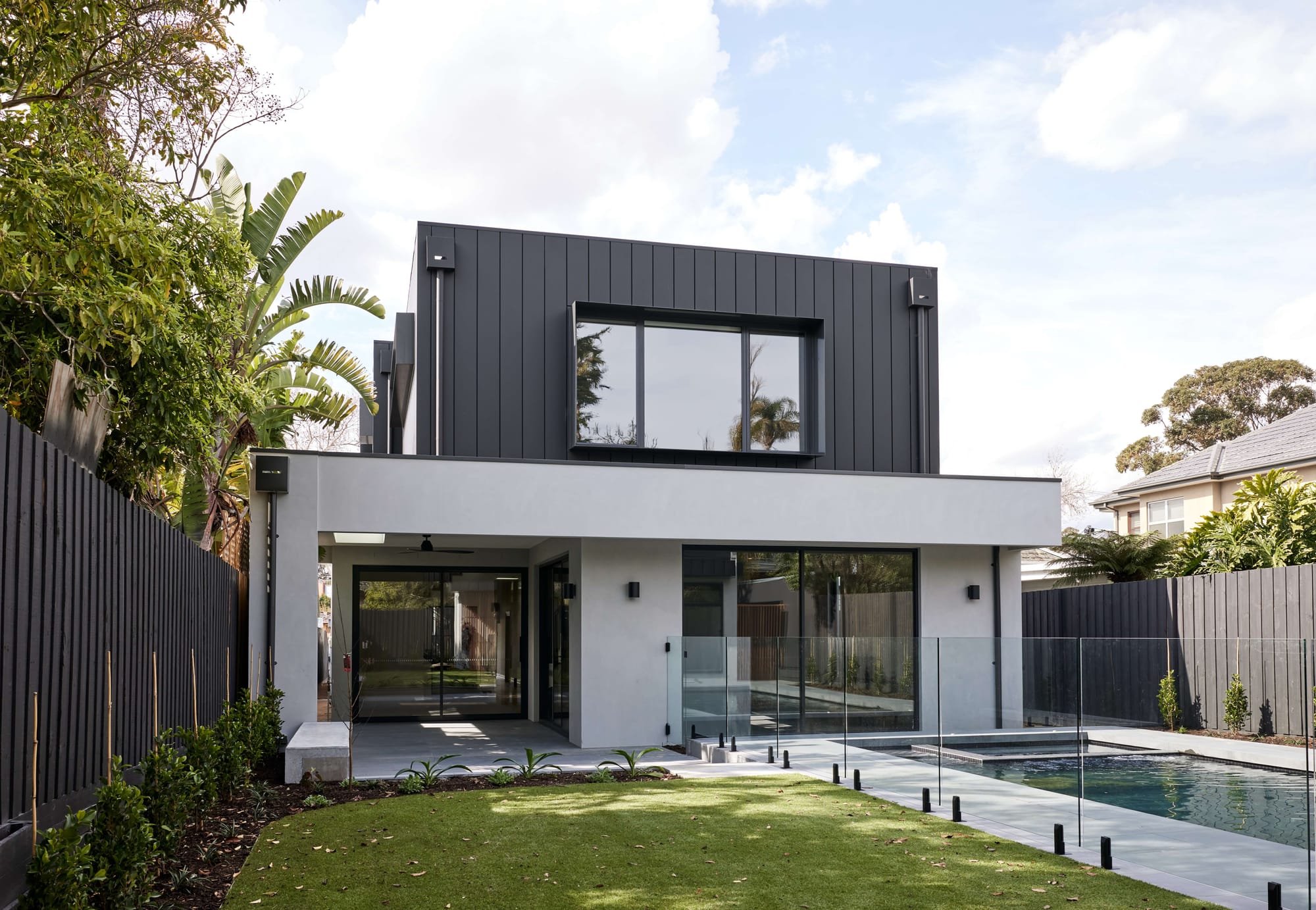 A dark timber-clad house with a corrugated metal gable roof, glimpsed through mature trees behind a pale timber-slat fence.