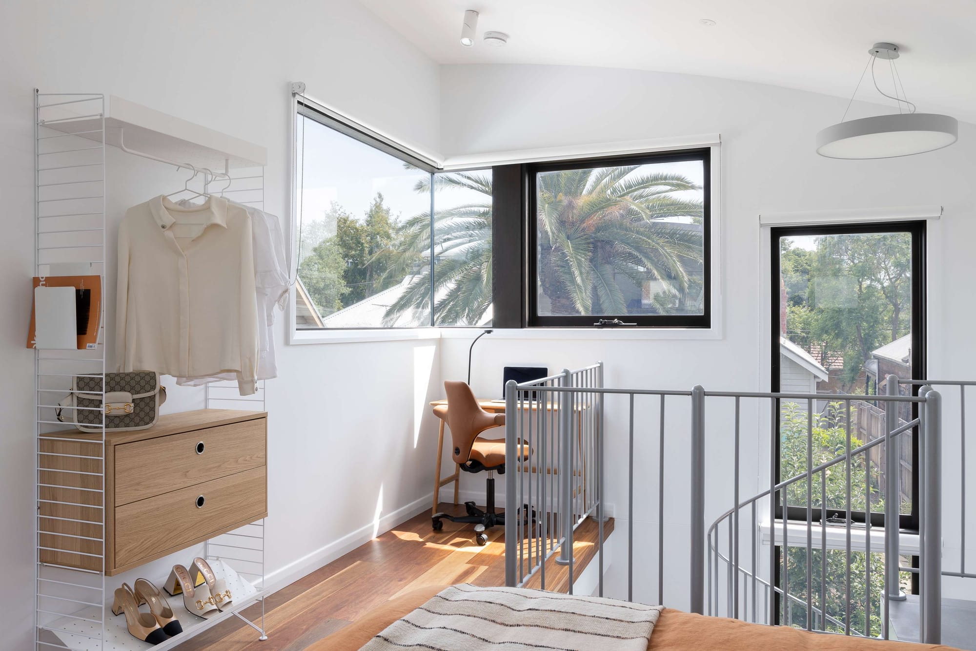 A mezzanine bedroom nook with a corner window framing palm trees, an open shelving wardrobe holding clothes and shoes, a timber desk with a leather chair, and a spiral staircase railing.