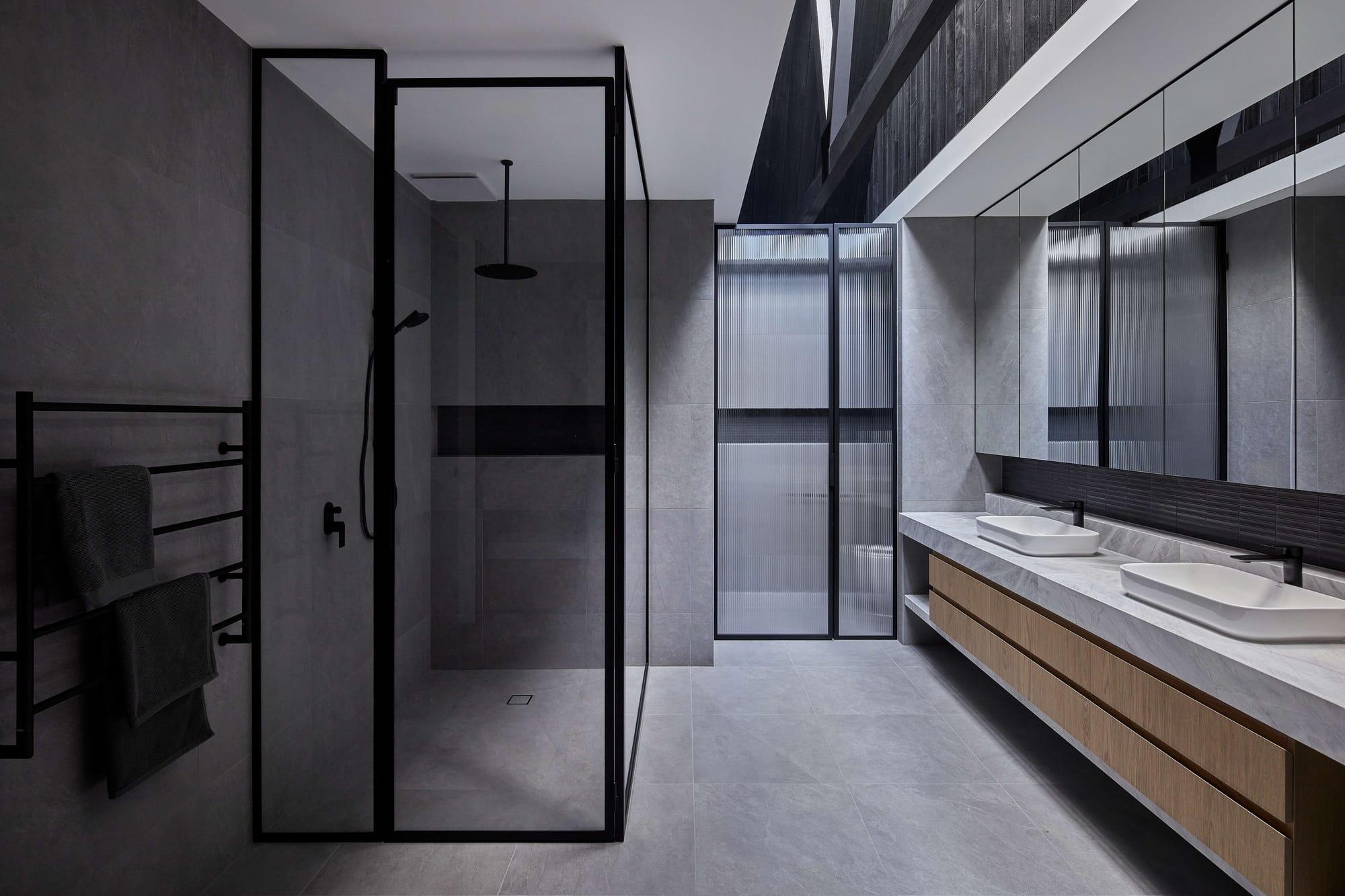 Contemporary bathroom with grey stone-look tiles, black-framed glass shower, double timber vanity, mirrored cabinetry and fluted glass privacy screens.