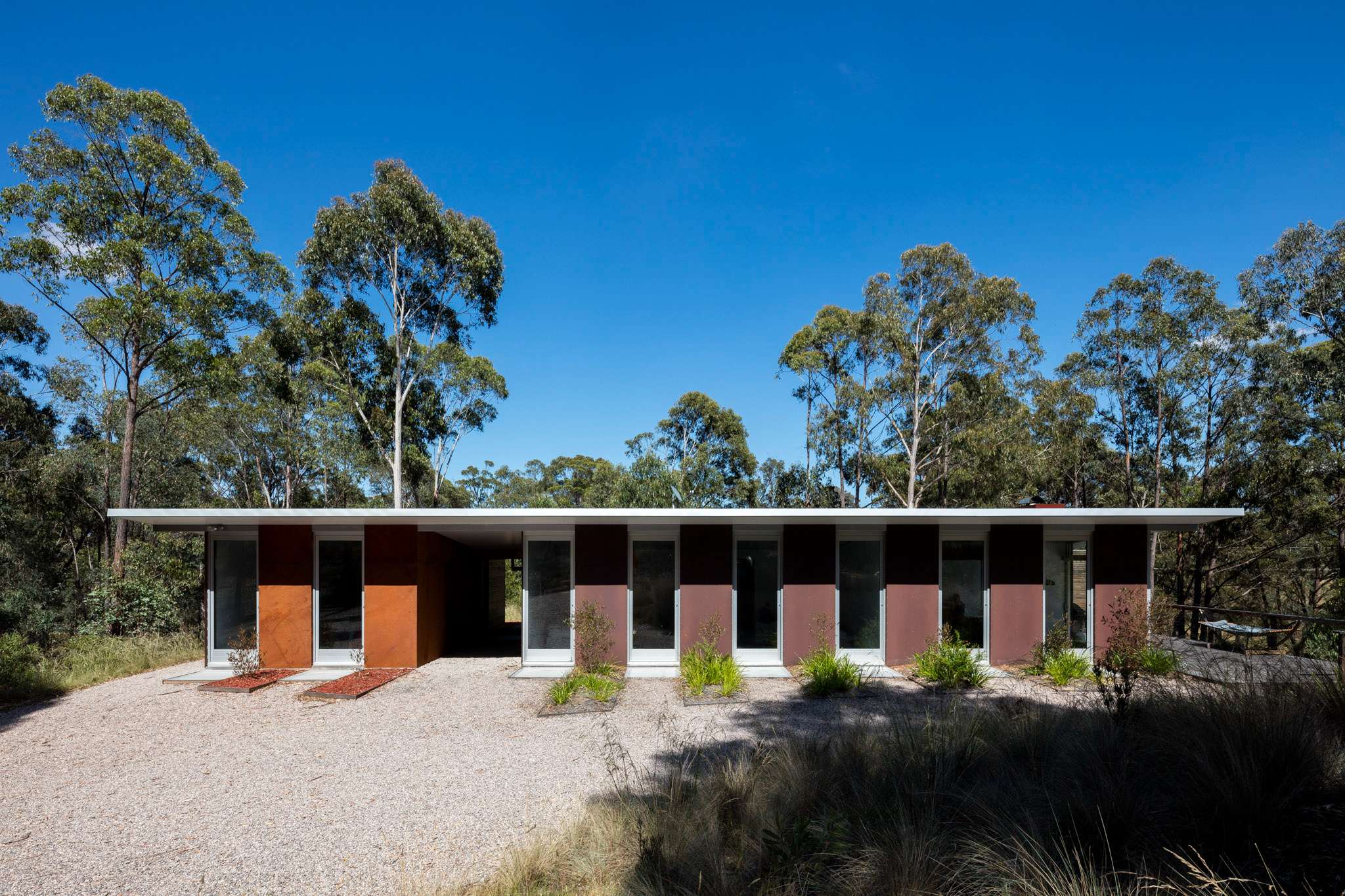 Single storey rural home with a long flat skillion roof, alternating rust orange and dark cladding panels, full height glazed doors along the facade, and native grasses and gravel landscaping to the front.