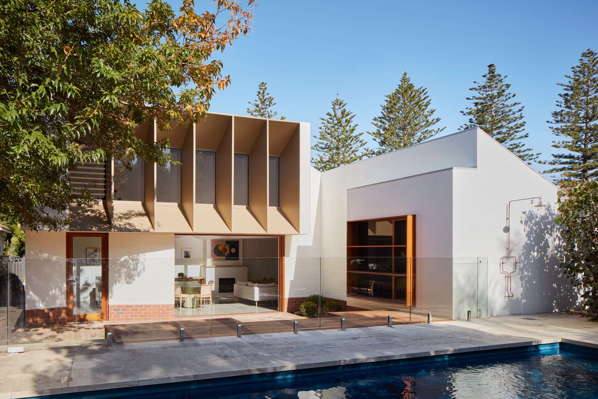 A white rendered beach house extension with an angled timber-lined upper bay window, opening through glass doors to a living room, beside a pool and Norfolk pines.