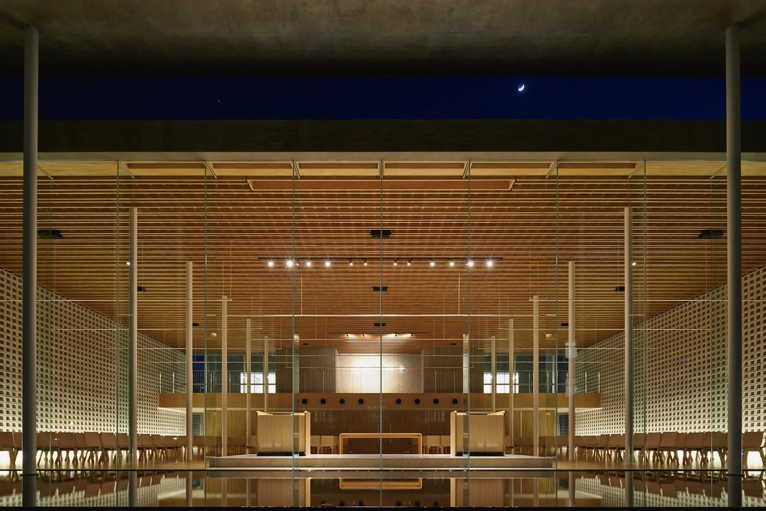 Symmetrical night view through a glazed façade into a timber-lined interior with fine vertical supports, patterned screens and reflective water in the foreground.