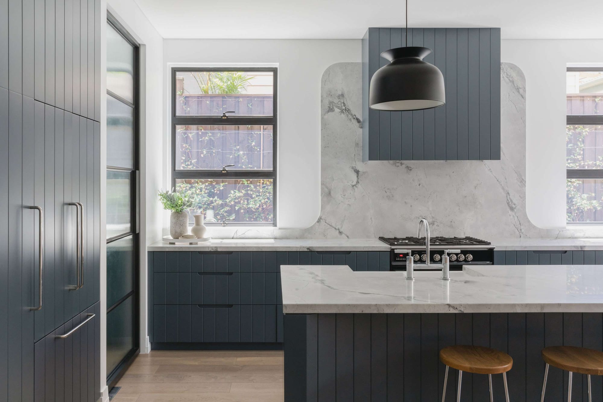 Kitchen with dark blue vertical panelled cabinetry, a curved marble splashback behind the cooktop, a black dome pendant light and a marble waterfall island with timber stools.
