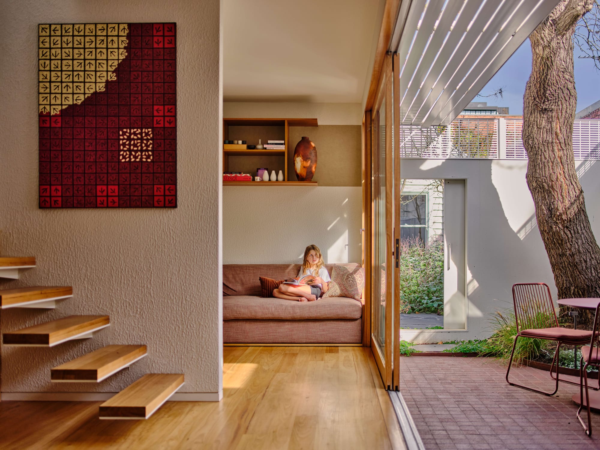 Girl reading on a sofa in a sunlit living nook, with a floating timber staircase and geometric wall tapestry, opening onto a courtyard with a mature tree through timber-framed sliding doors and an adjustable louvred awning above.