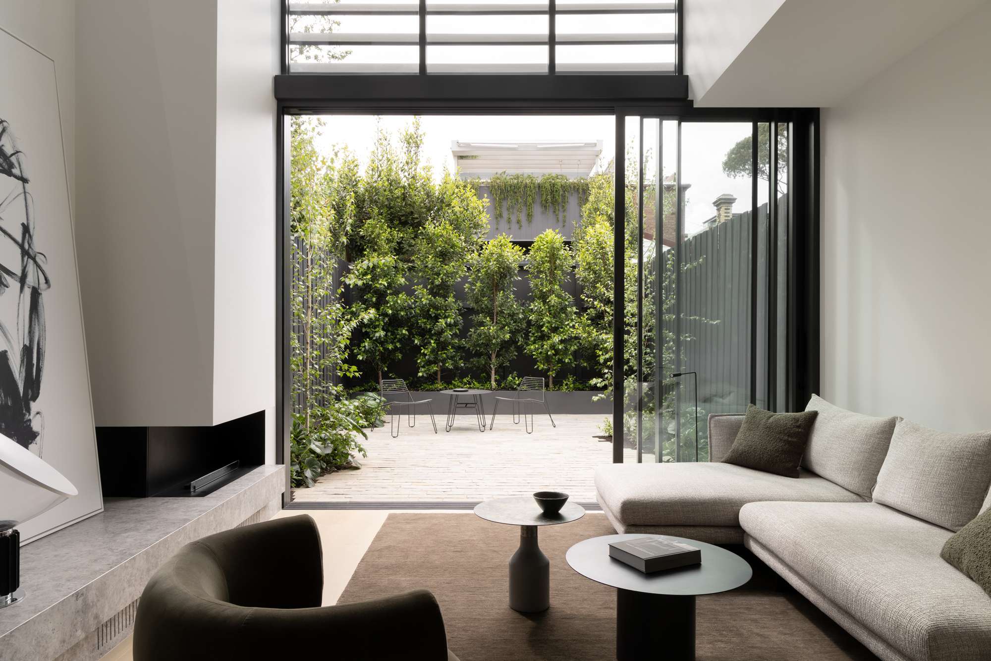 Living room with a corner fireplace, grey sectional sofa, and full-height sliding glass doors opening onto a paved courtyard with wire chairs and a lush green hedge screen.